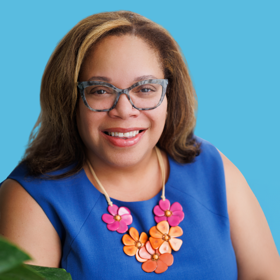 A woman with glasses and a colorful floral necklace smiling at the camera, wearing a blue sleeveless top, against a blue background.