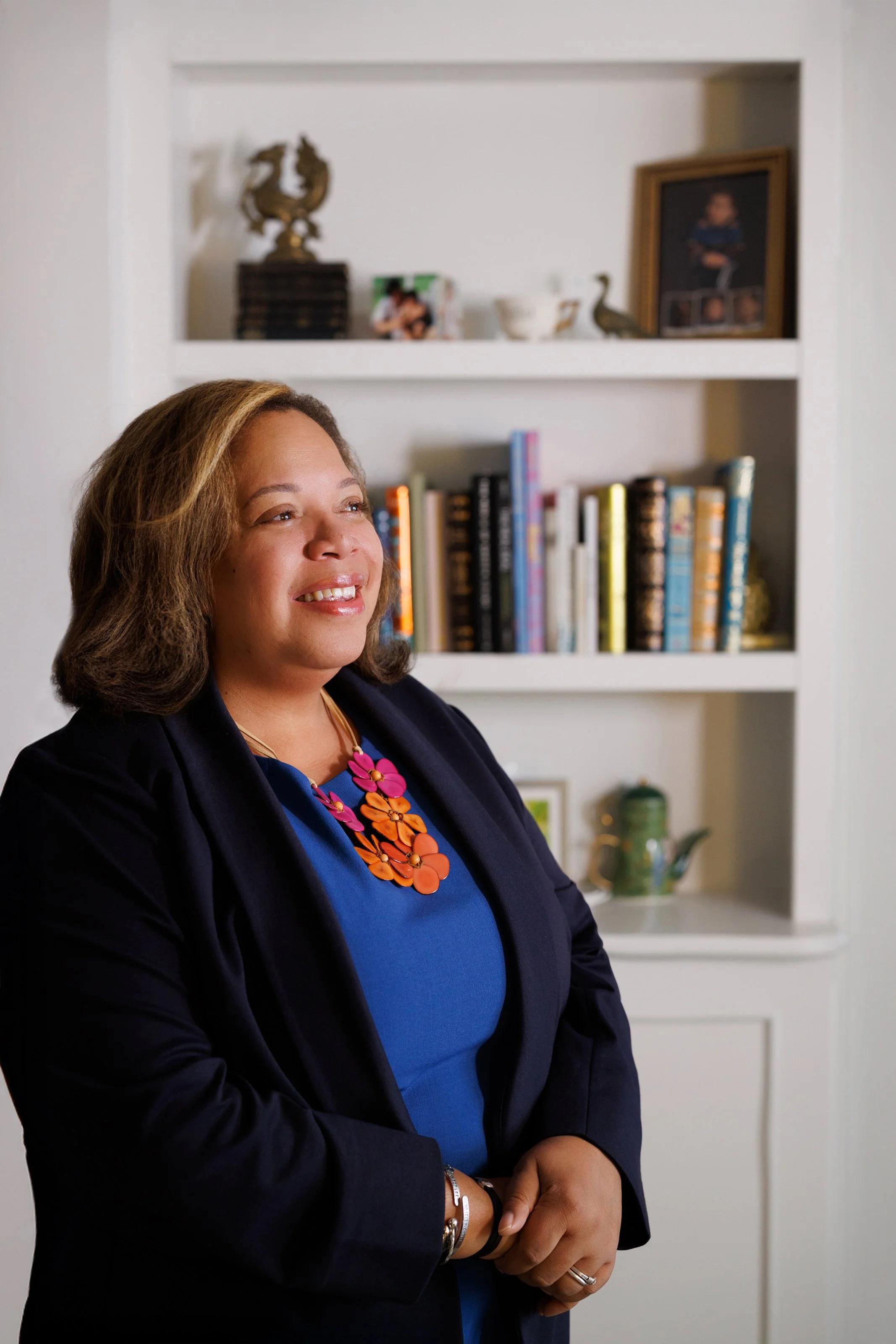 A smiling woman with medium-length brown hair, wearing a navy blazer, a blue top, and a colorful statement necklace. She stands with her hands crossed in front of her, in front of a white wall with built-in shelves filled with books, decorative objects, a framed photograph, and a small sculpture.