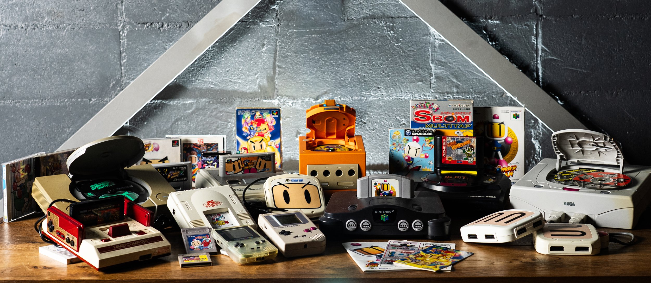 Collection of vintage and modern video game consoles, games, and accessories displayed on a wooden surface with a metal wall backdrop.