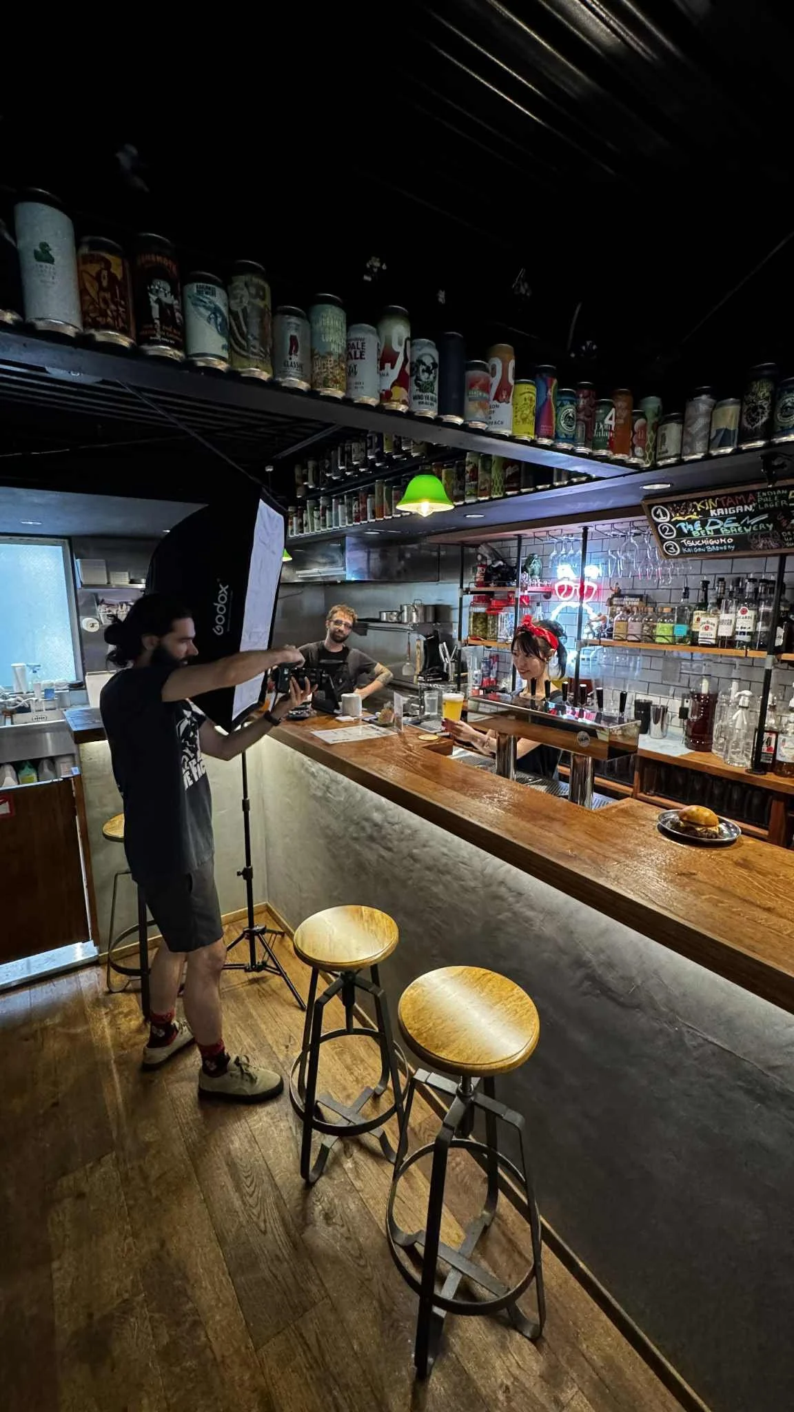 A person filming a woman behind a bar in a cozy, dimly lit bar with bar stools and shelves of liquor bottles.