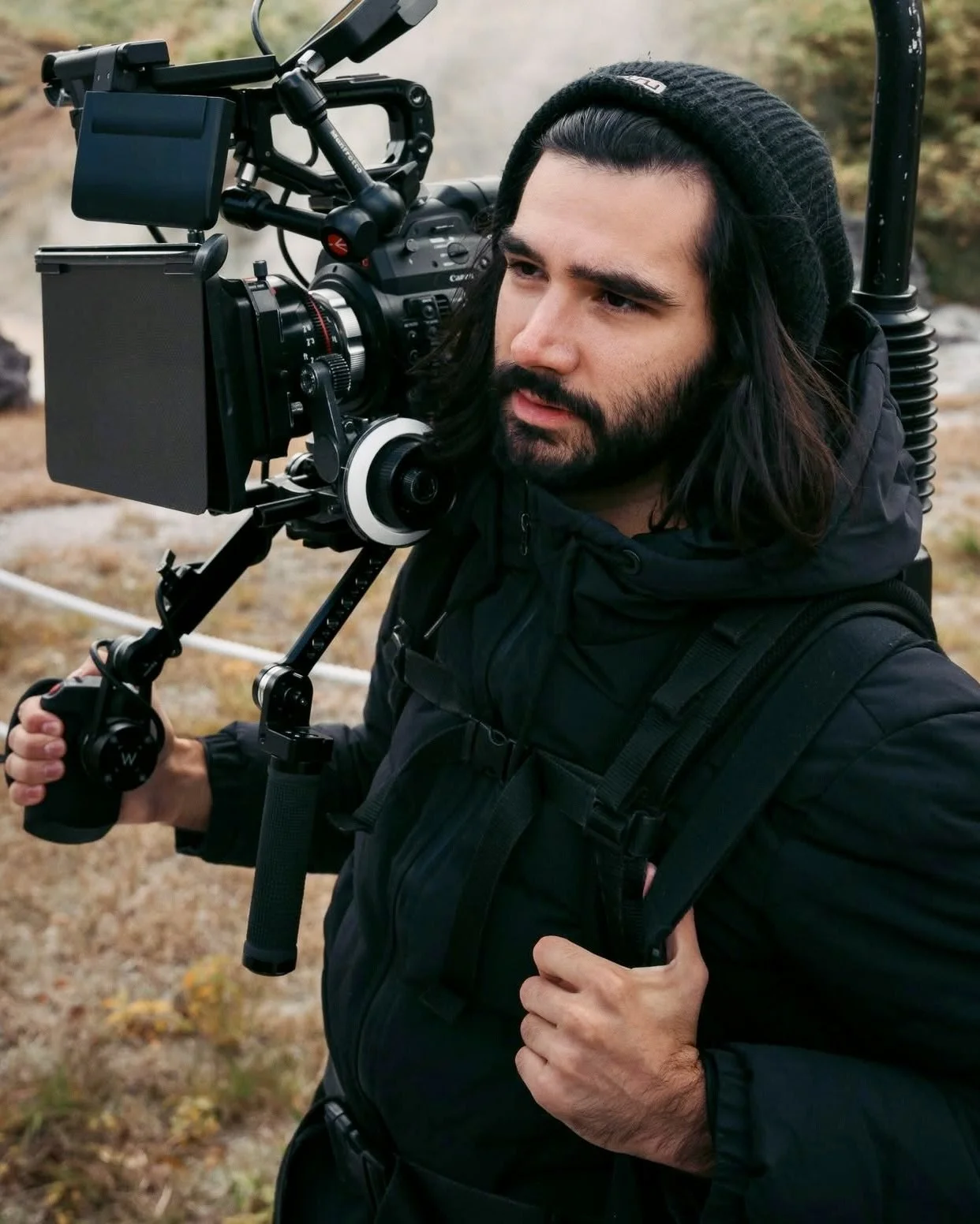 A man with long black hair and a beard operating a professional camera on a shoulder rig outdoors.