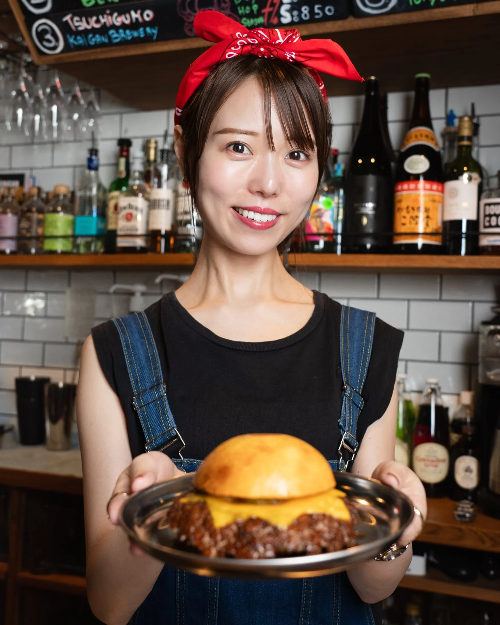A smiling woman with short brown hair, wearing a red bandana and denim dress, holds a plate of food towards the camera in a restaurant setting with liquor bottles and a menu board in the background.