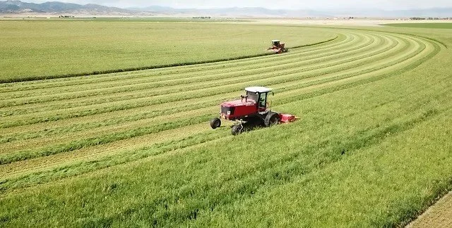 Two red tractors working on a lush green agricultural field, creating curved lines in the grass with distant mountains in the background.