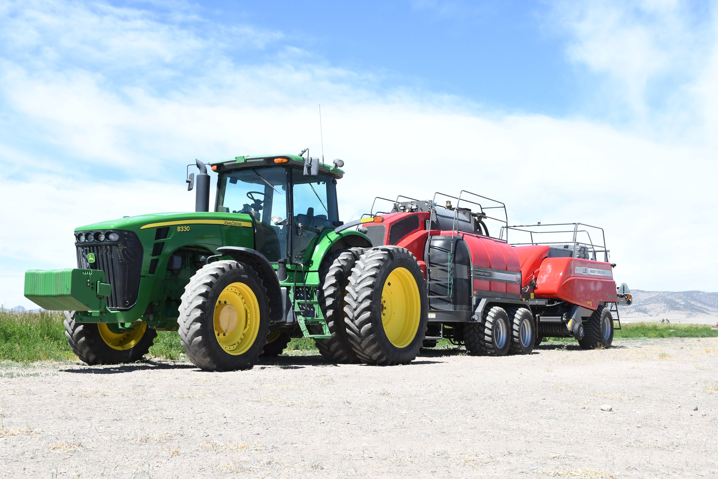 A green John Deere tractor pulling a red Massey Ferguson fertilizer or sprayer tank in an open field on a bright, partly cloudy day.