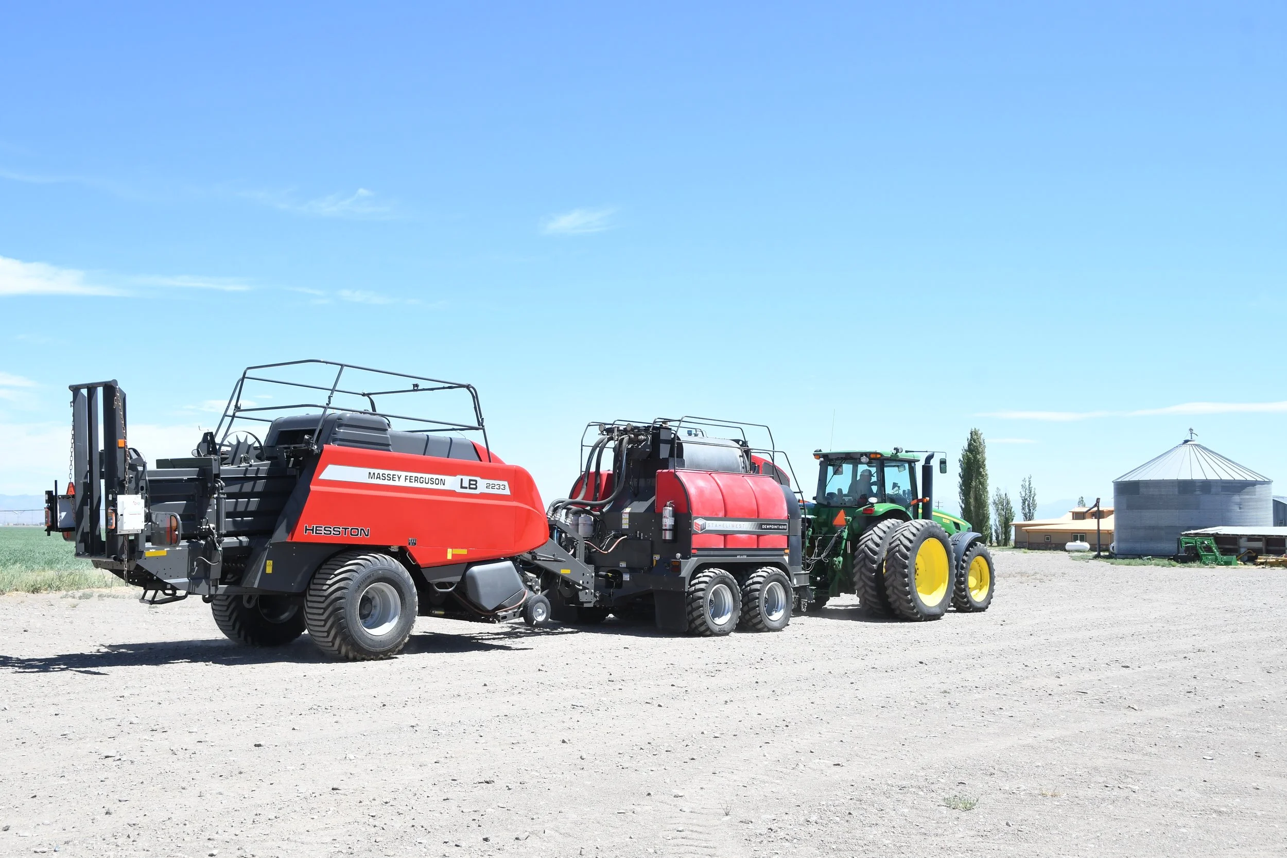 Large agriculture machinery in a field, including a Massey Ferguson Hesston baler, a red sprayer, and a green tractor, with farm buildings and silos in the background under a clear blue sky.
