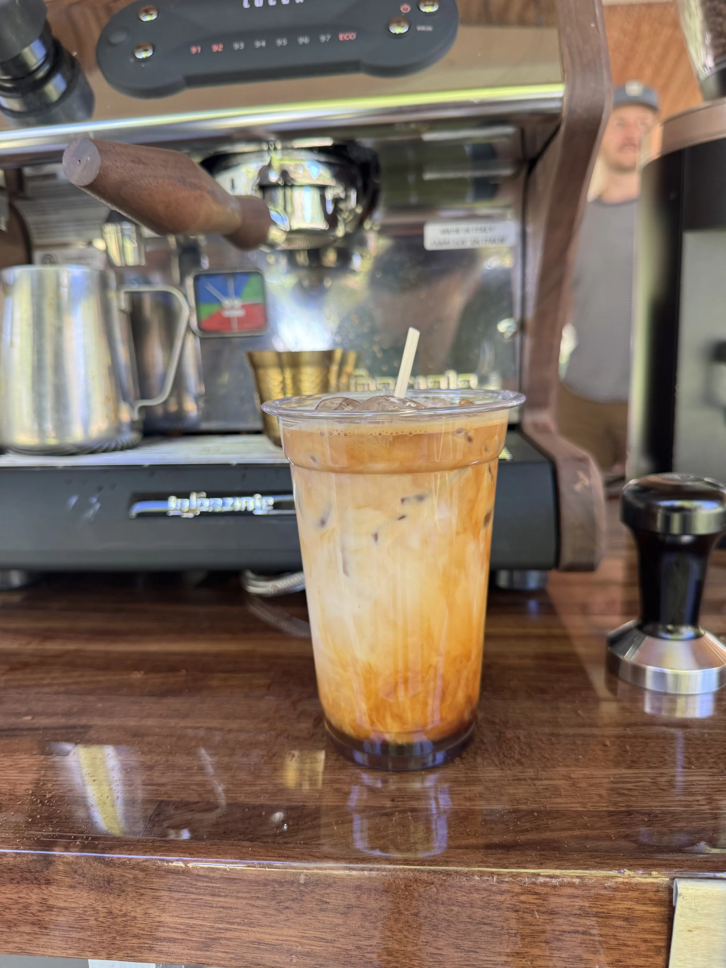 A plastic cup filled with iced coffee or iced tea with a straw on a wooden table, with a coffee machine, coffee scoop, and other coffee-related items in the background.