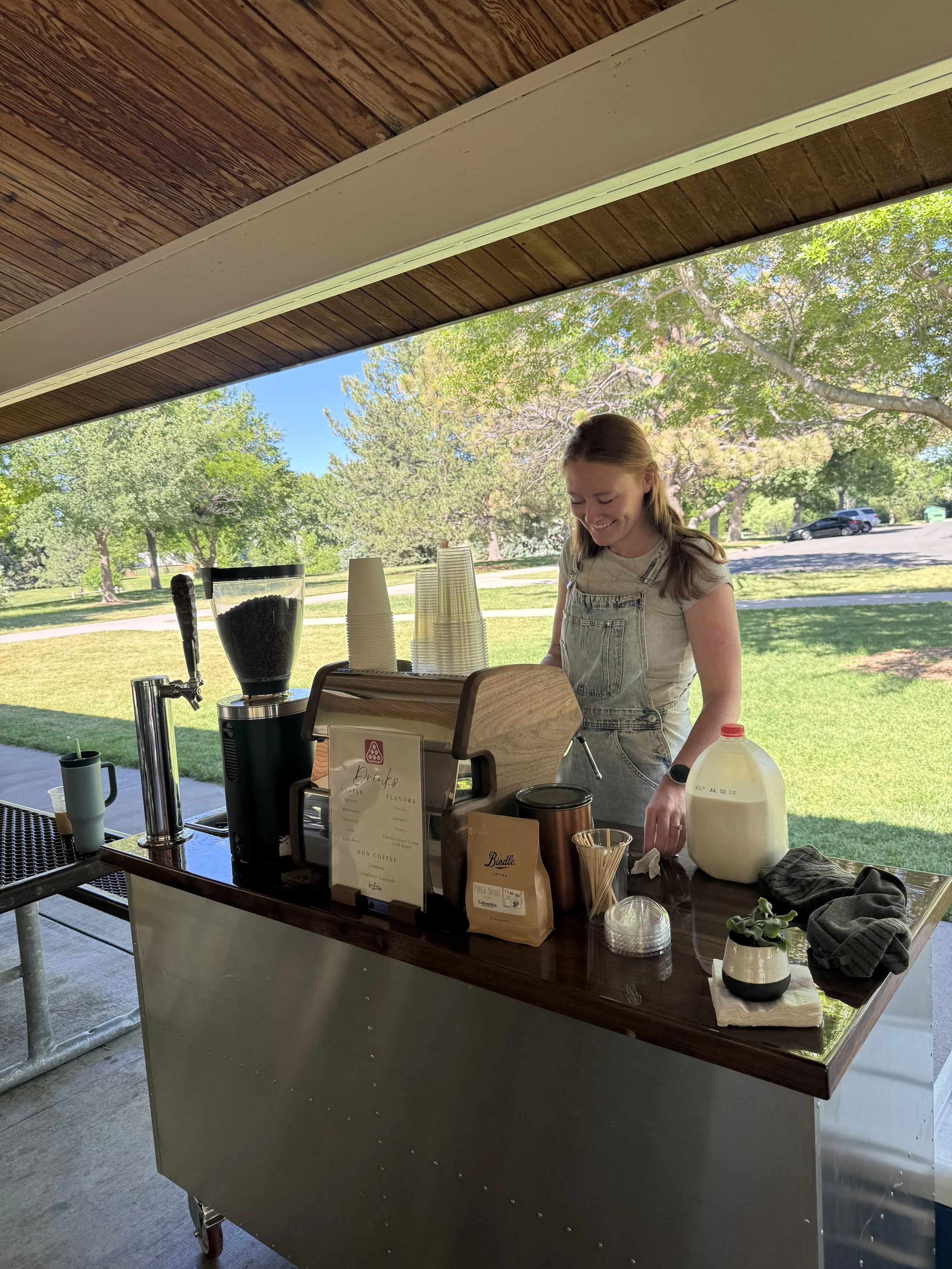 Smiling woman in casual clothing preparing coffee at an outdoor coffee stand with trees and parking lot in the background.