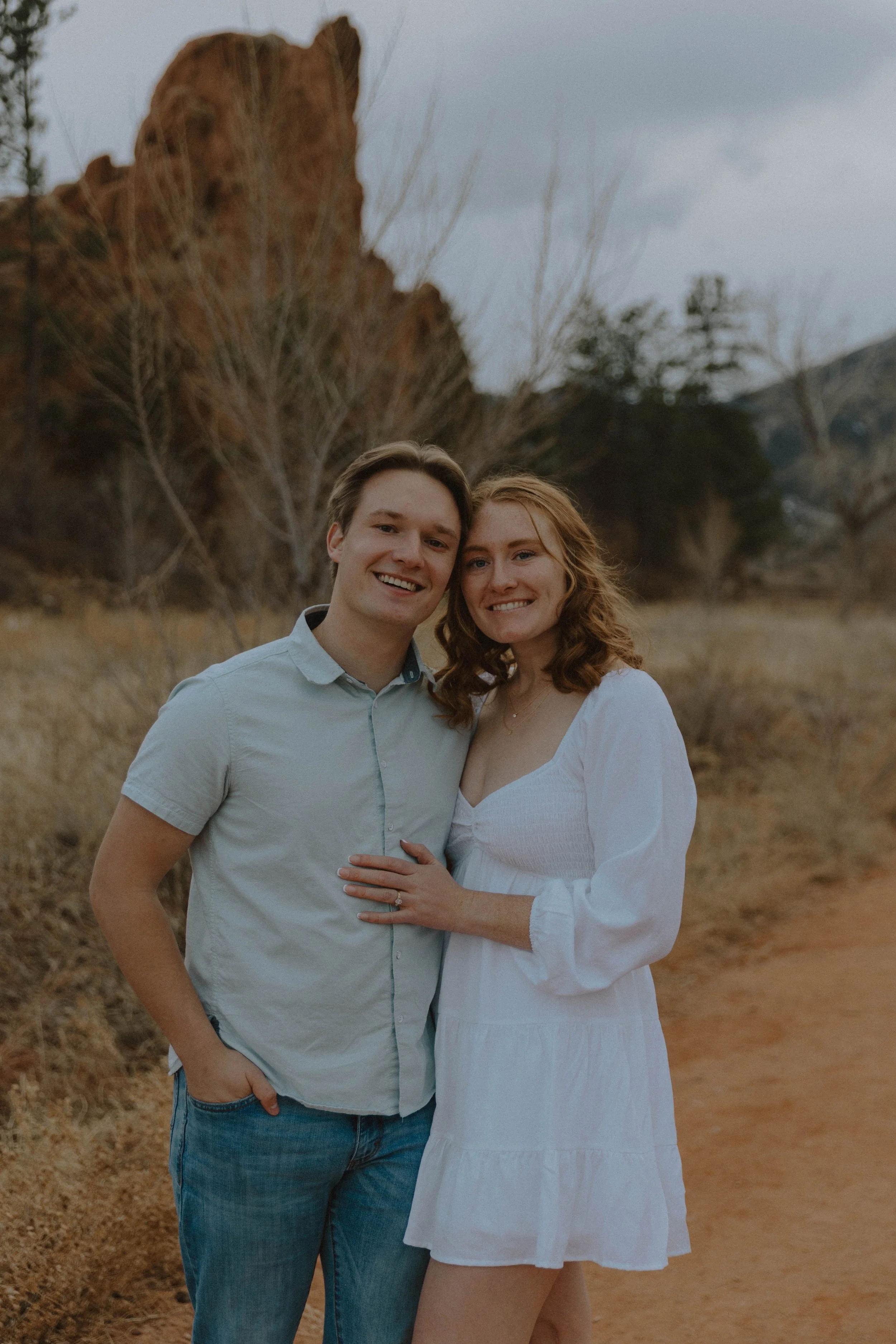 A smiling young man and woman standing outdoors in a rural area with dry grass and trees in the background, looking at the camera.