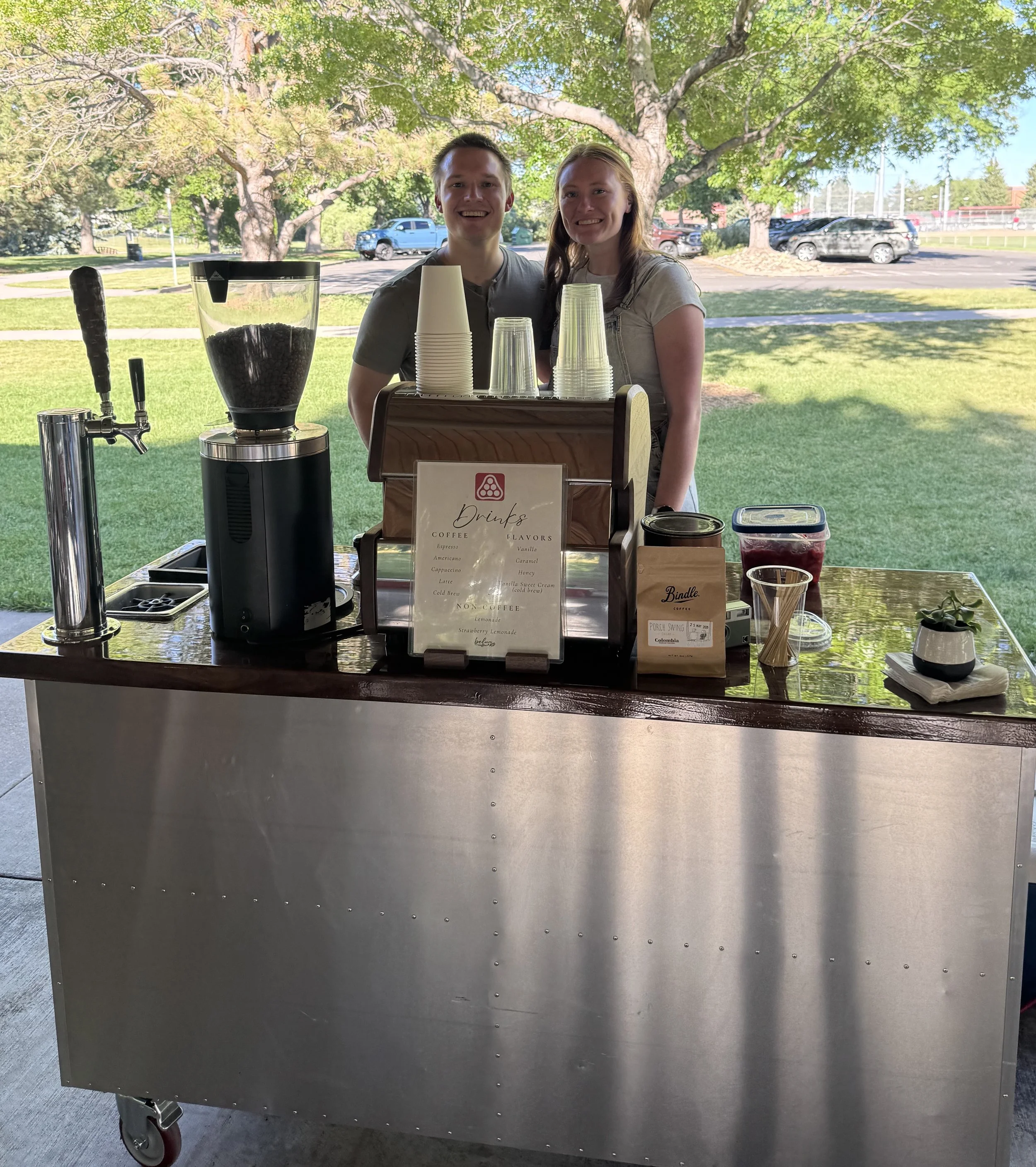 A metal cart with drinks and coffee supplies seen from the front, outdoors on a sunny day with trees and parked cars in the background.