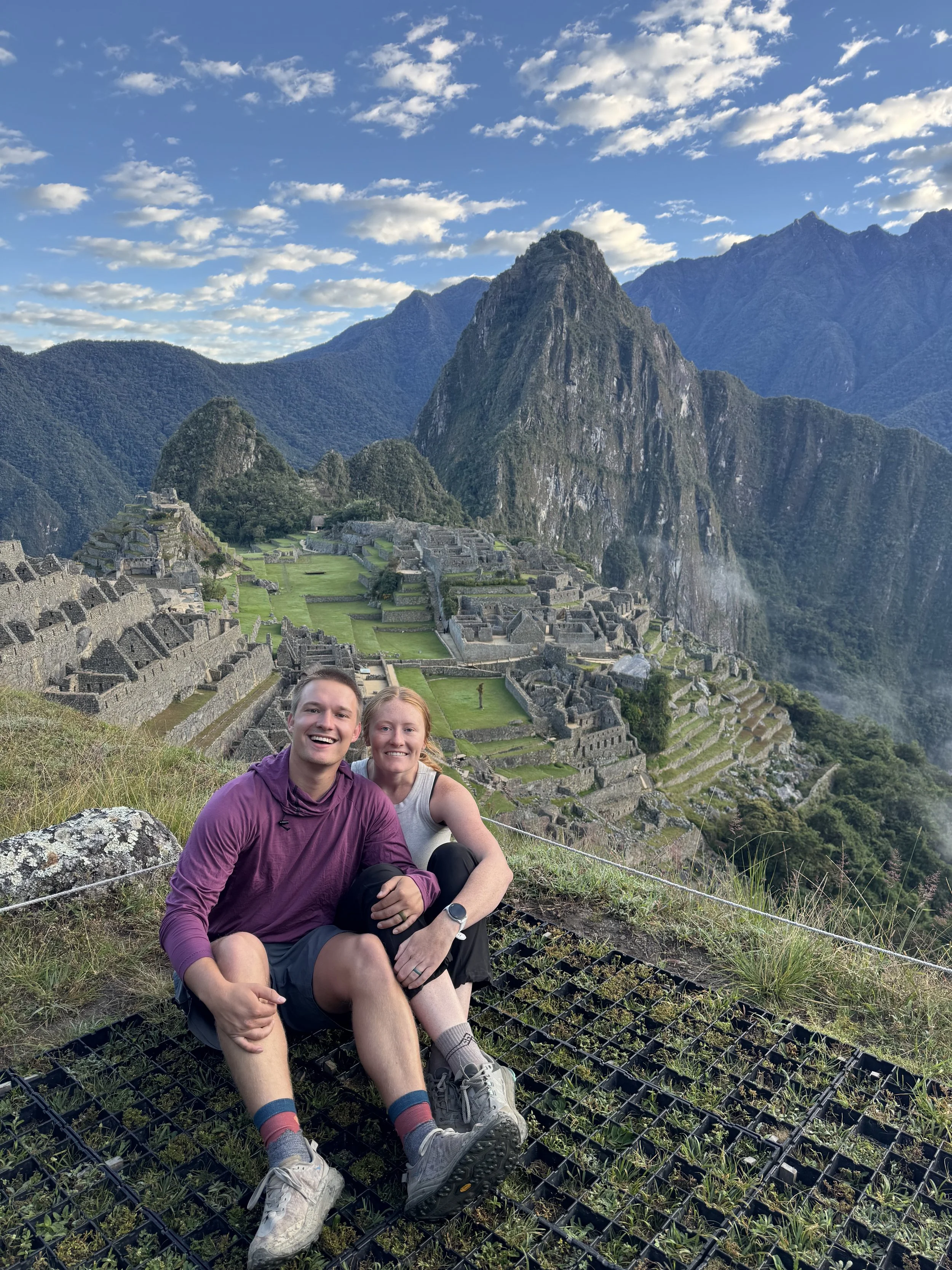 Two smiling hikers, a man and a woman, sitting on the ground with Machu Picchu in the background, surrounded by mountains and a partly cloudy sky.
