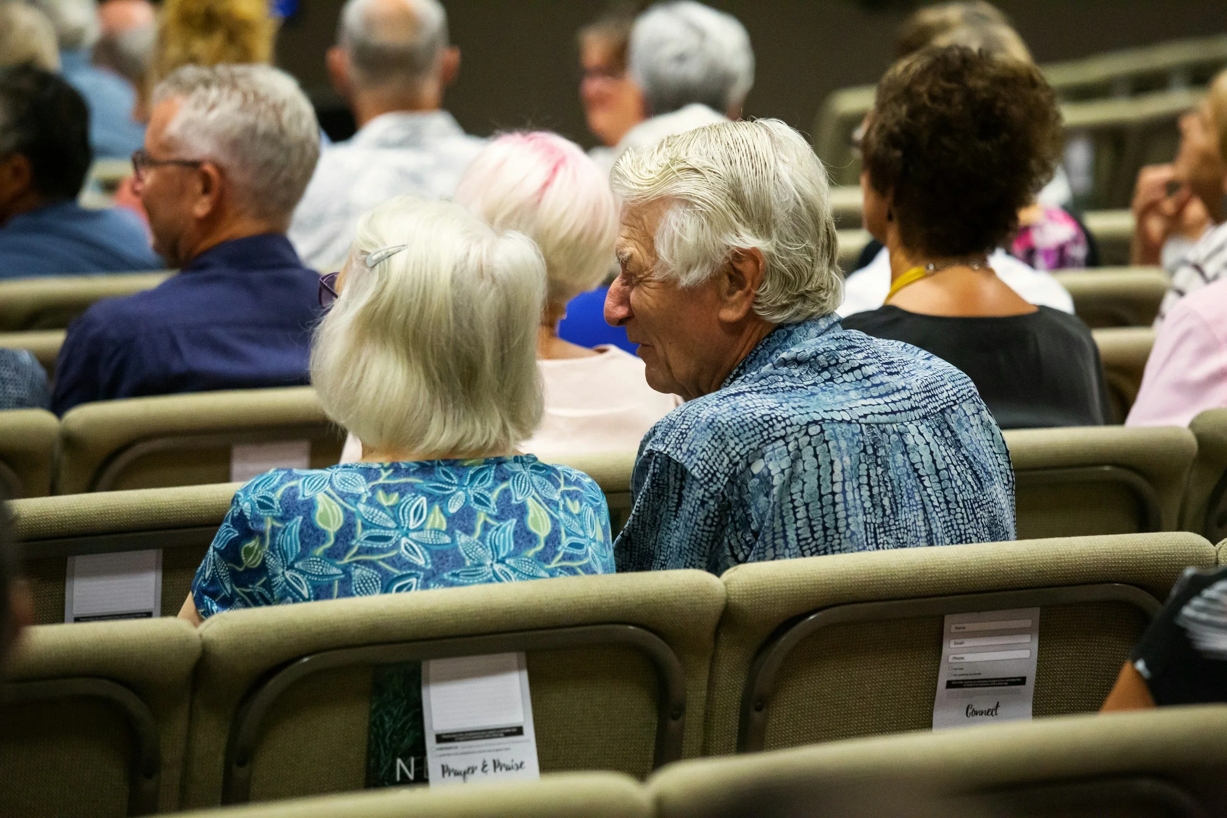 People sitting in an auditorium, some engaged in conversation, with chairs labeled for different activities and interests. The setting appears to be a conference or seminar.