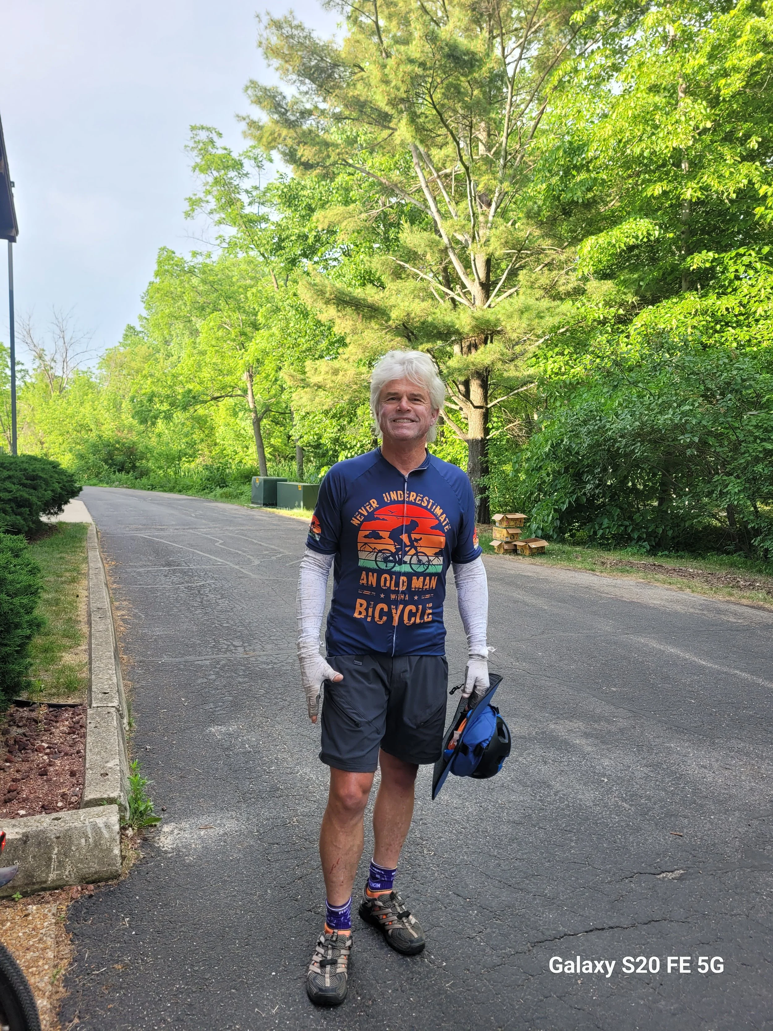 Dave Hodges holding a helmet, standing on a paved road surrounded by green trees on a bright day.