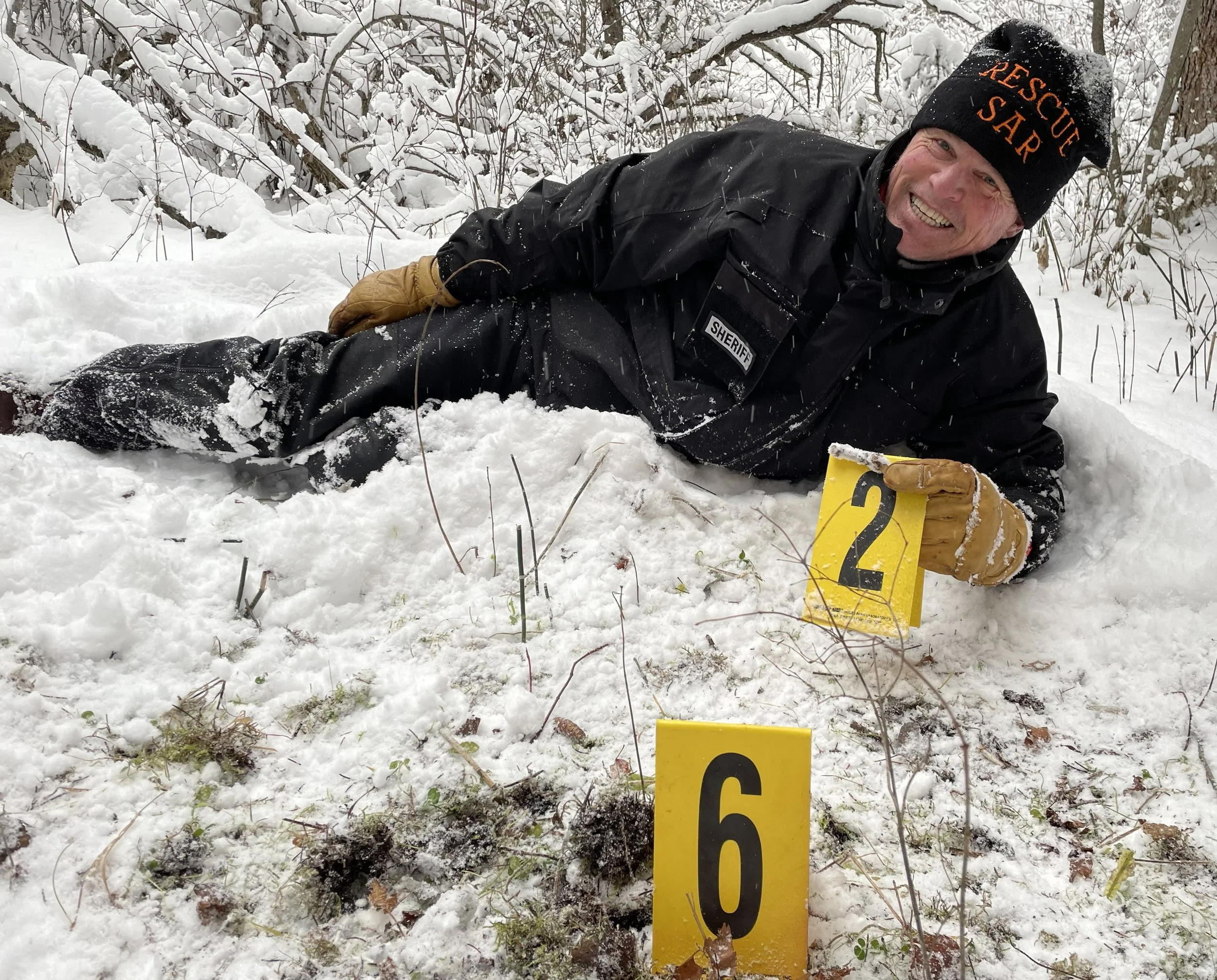 Dave Hodges lying in snow with a yellow evidence marker labeled 6 nearby, surrounded by snow-covered branches and bushes.