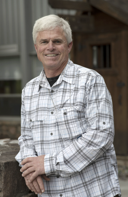 A smiling Dave Hodges, wearing a light plaid shirt, standing outdoors in front of a rustic wooden building.
