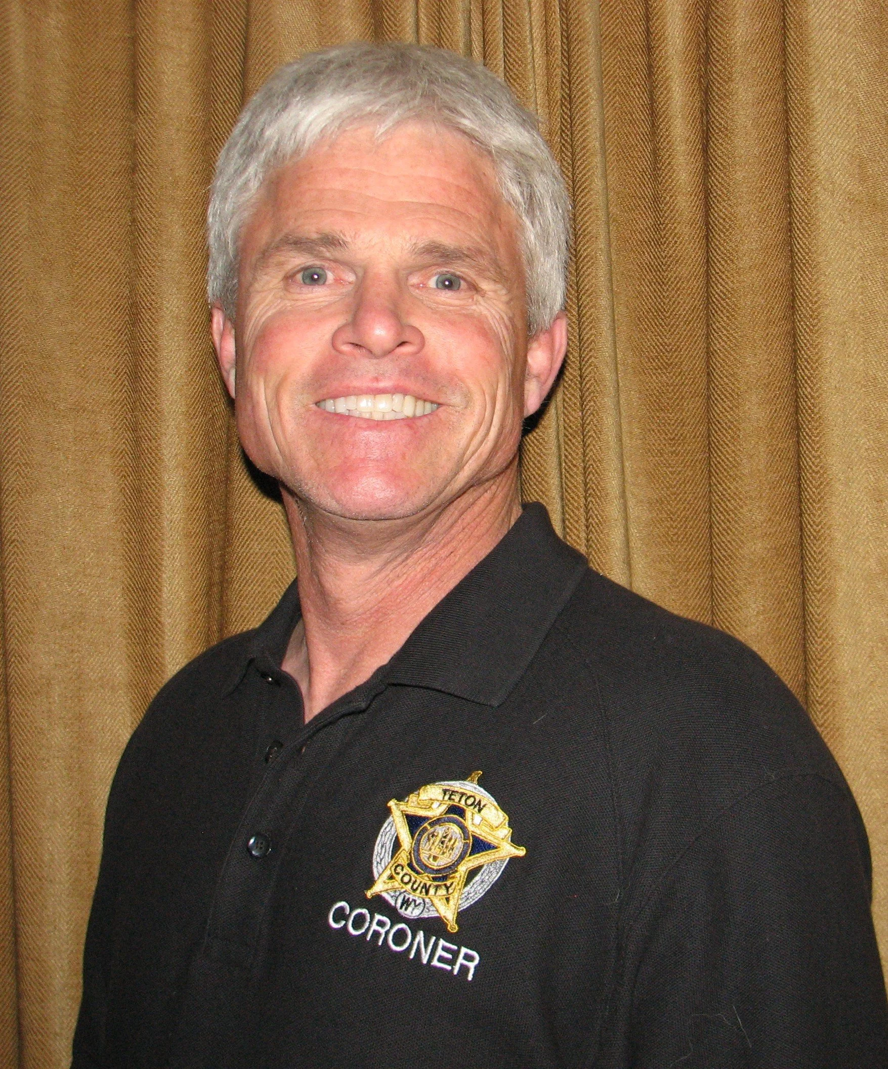 Close-up of Dave Hodges wearing a black shirt with a Sheriff badge that says 'Teton County Coroner' standing in front of a gold curtain.