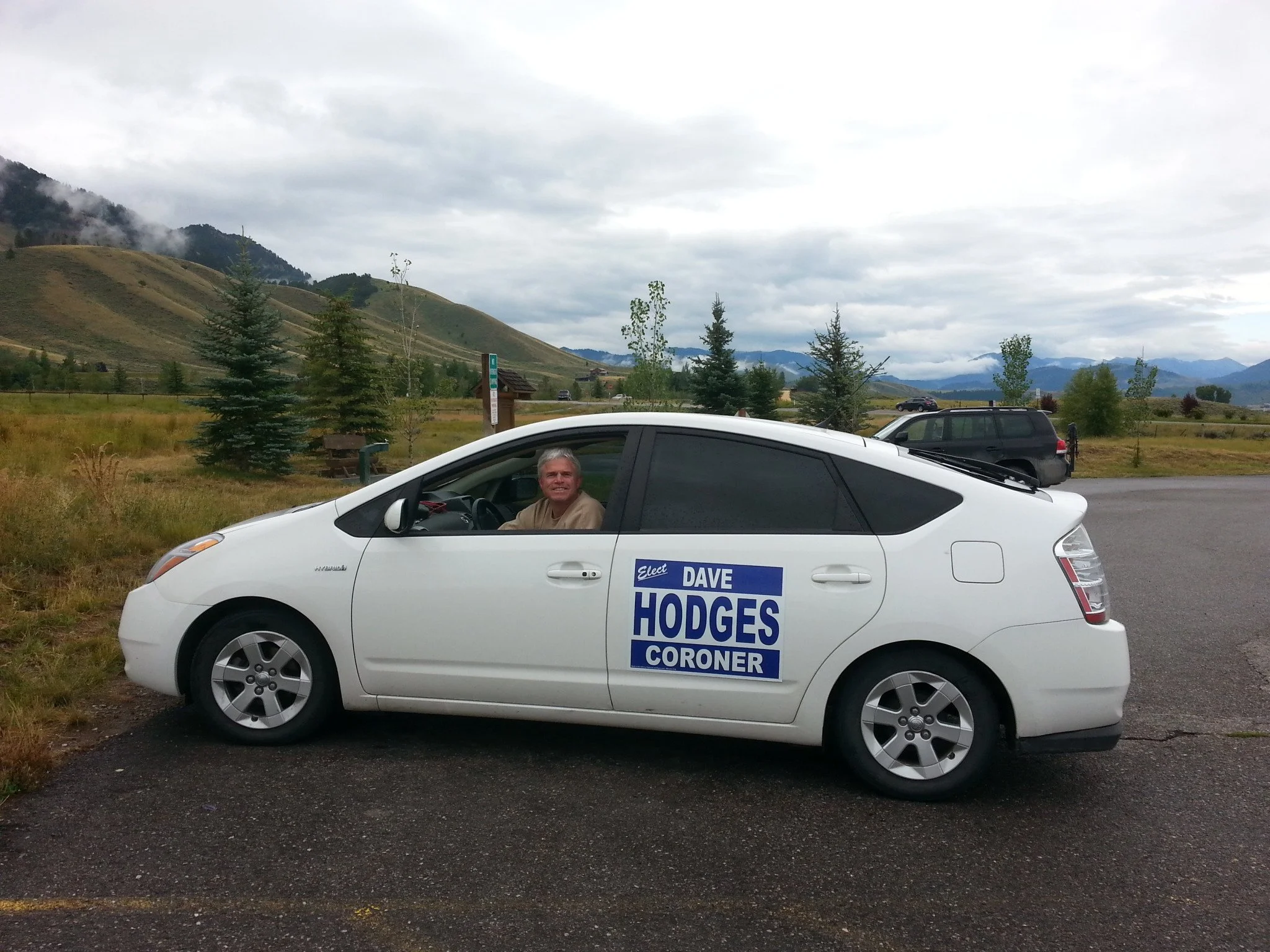 Dave Hodges in the driver's seat of a white car with a political campaign sign for Dave Hodges on the side door, with a scenic mountain landscape in the background.