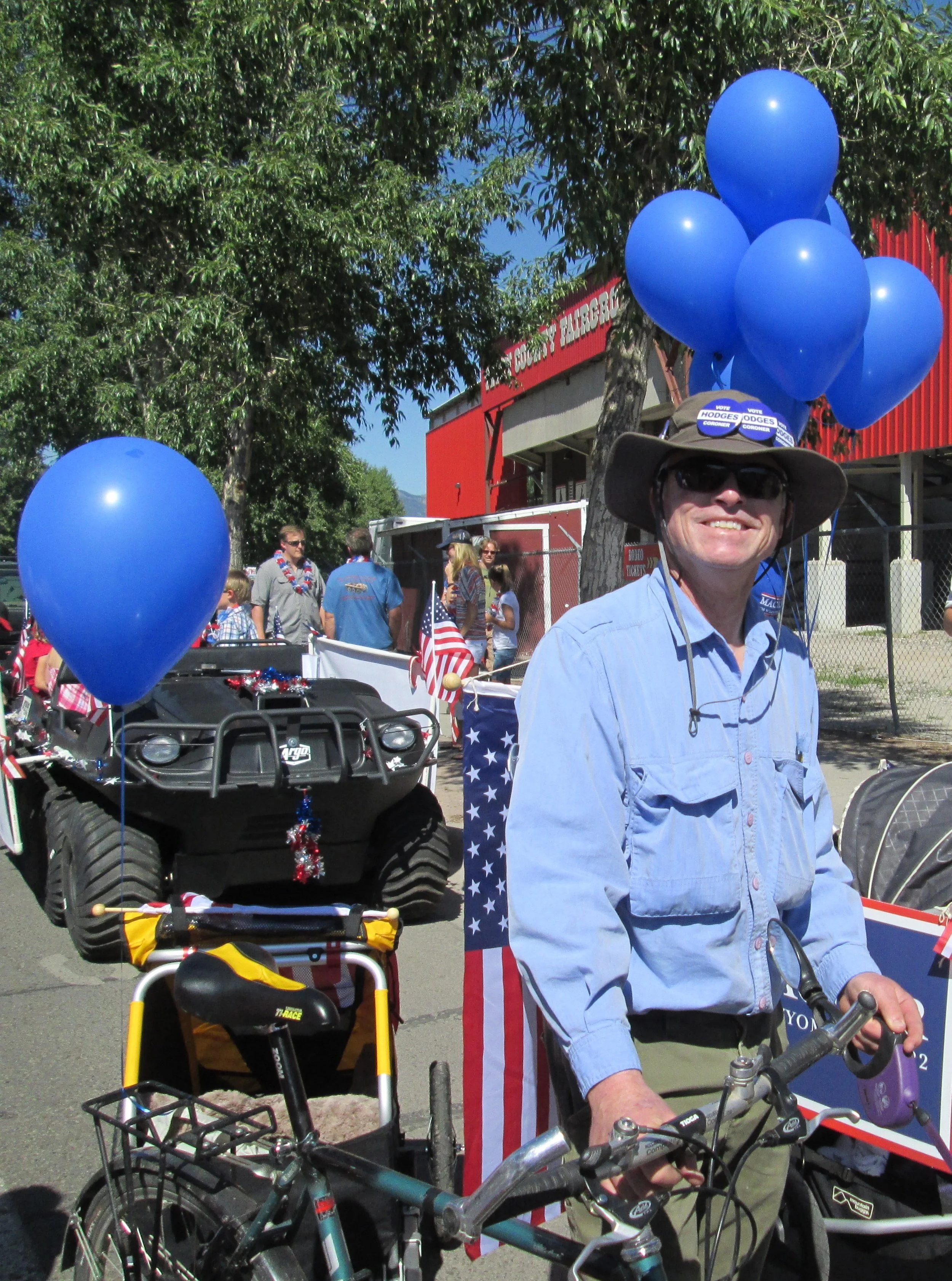 Dave Hodges riding a bicycle during a parade or celebration, with balloons and patriotic decorations in the background, including an American flag.