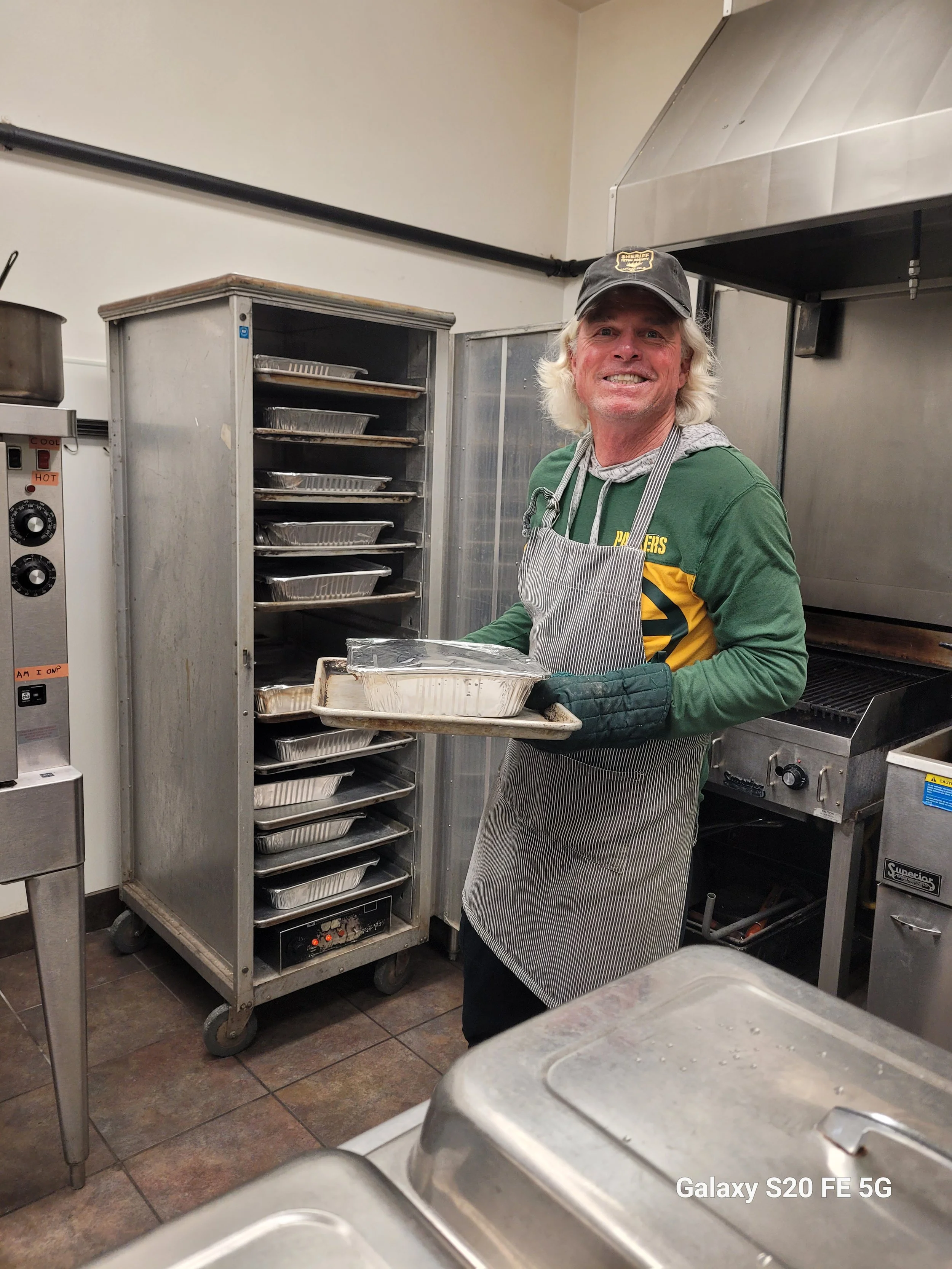 Dave Hodges a cap, apron, and gloves is standing in a commercial kitchen, holding a tray with aluminum foil-lined trays in front of an open industrial oven with multiple trays inside.