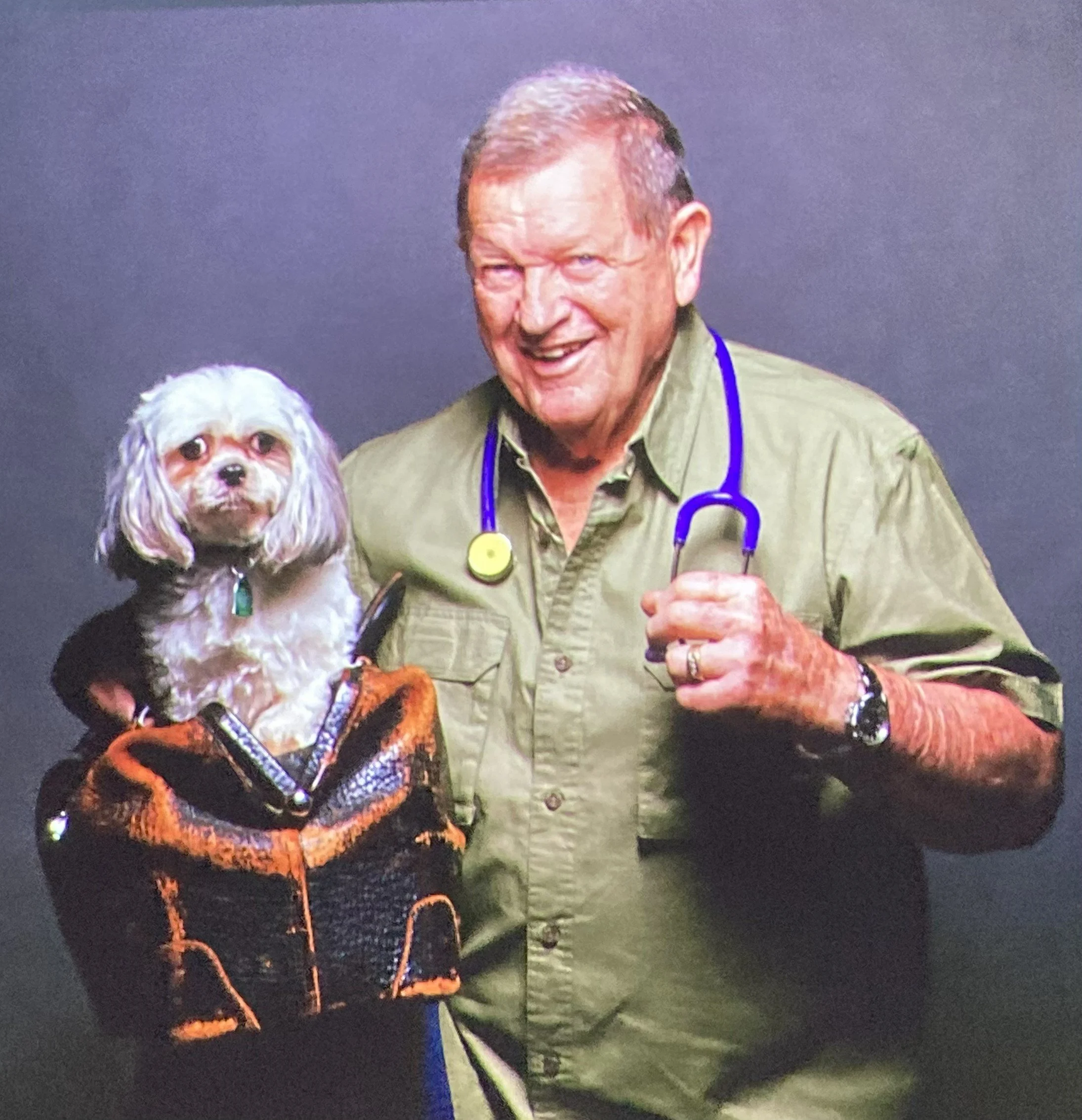 A man smiling while holding a glass and standing next to a dog wearing an orange vest in a dark setting.