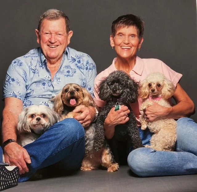 A smiling elderly couple sitting on the floor with four small dogs in front of a gray background.