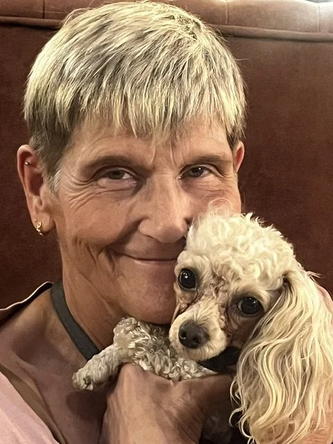 A woman holding a small, curly-haired dog close to her face, both looking at the camera inside a room with warm lighting.