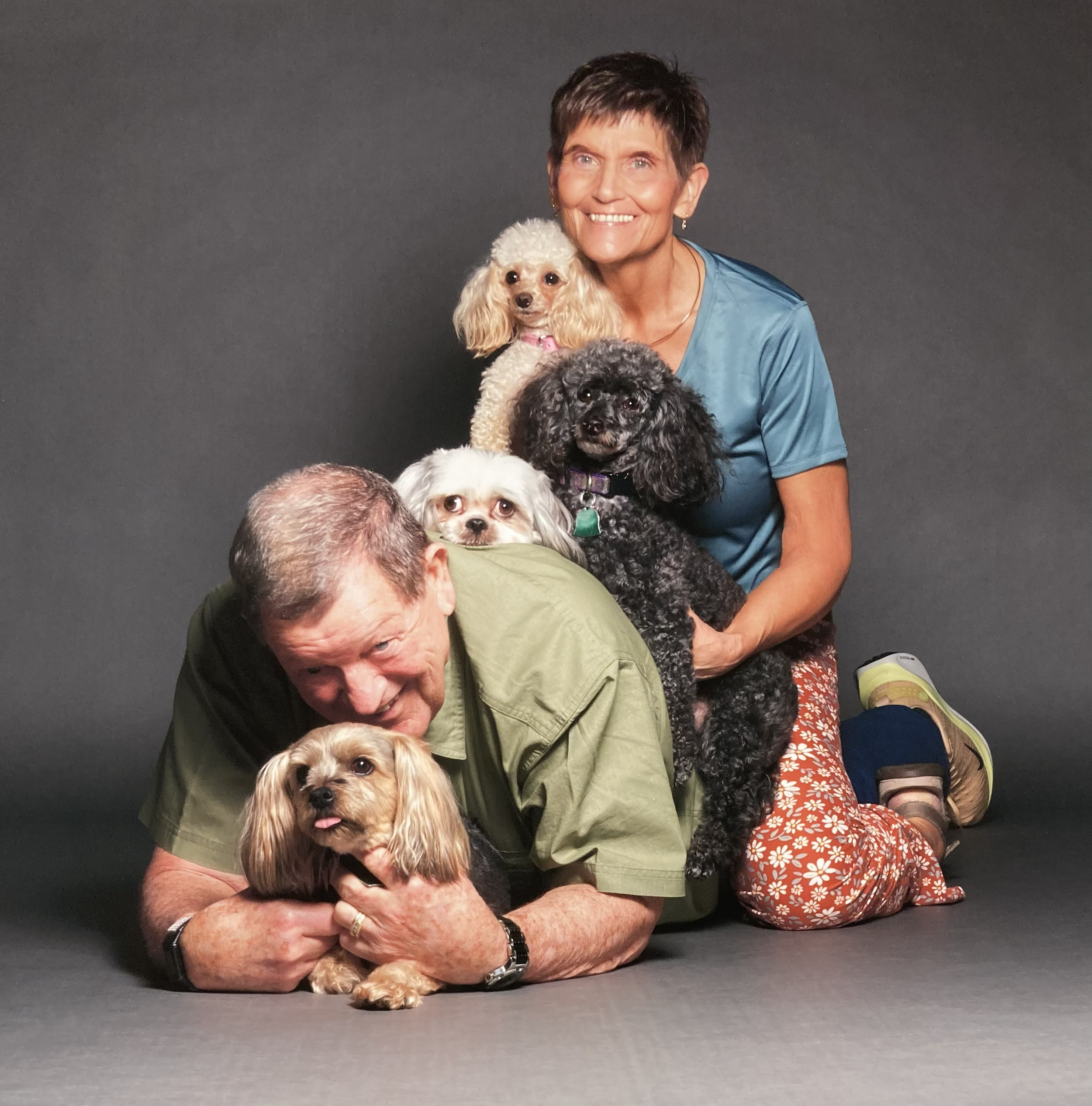 A smiling woman with short dark hair holding a light-colored curly-haired dog, along with other dogs, against a dark gray background, with a man bending down to cuddle another curly-haired dog in the foreground.