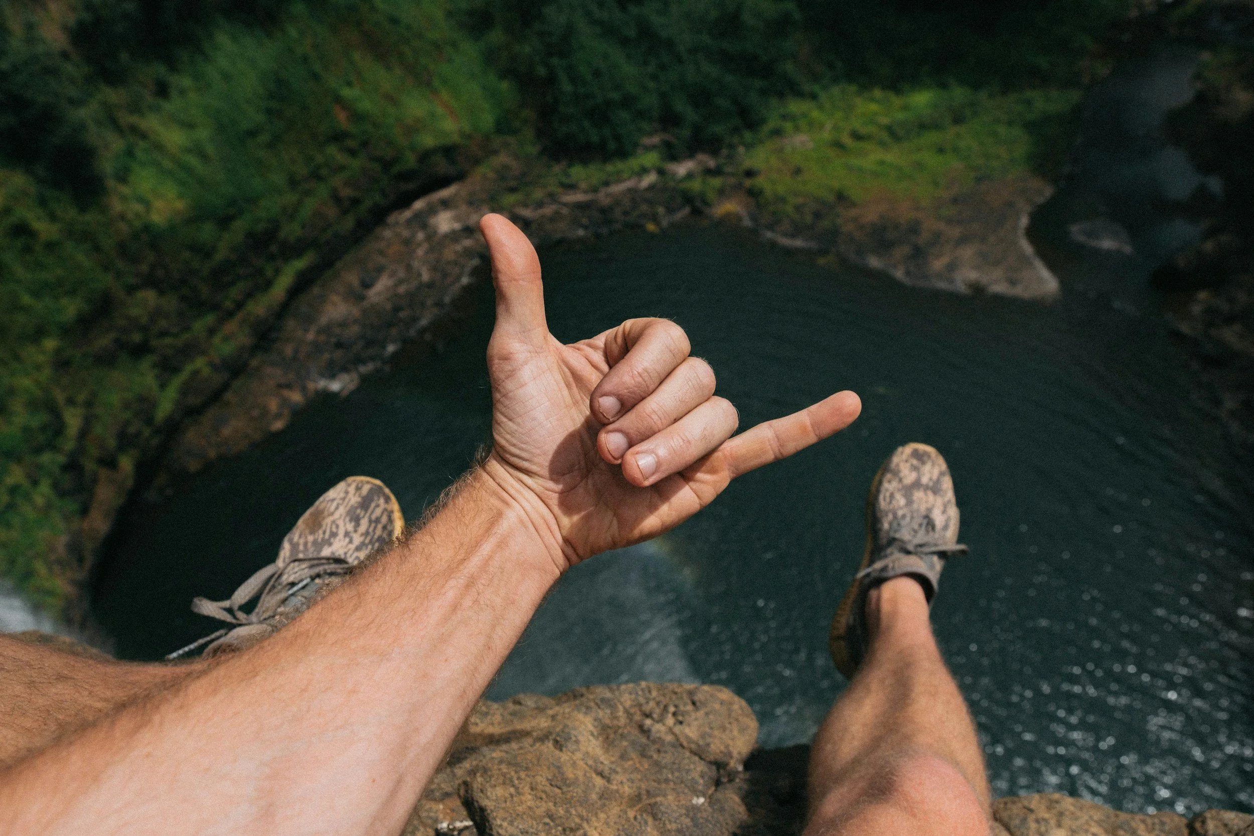 Person standing on a rocky ledge overlooking a body of water with lush green hills in the background, holding up one hand with the thumb, index, and pinky fingers extended.