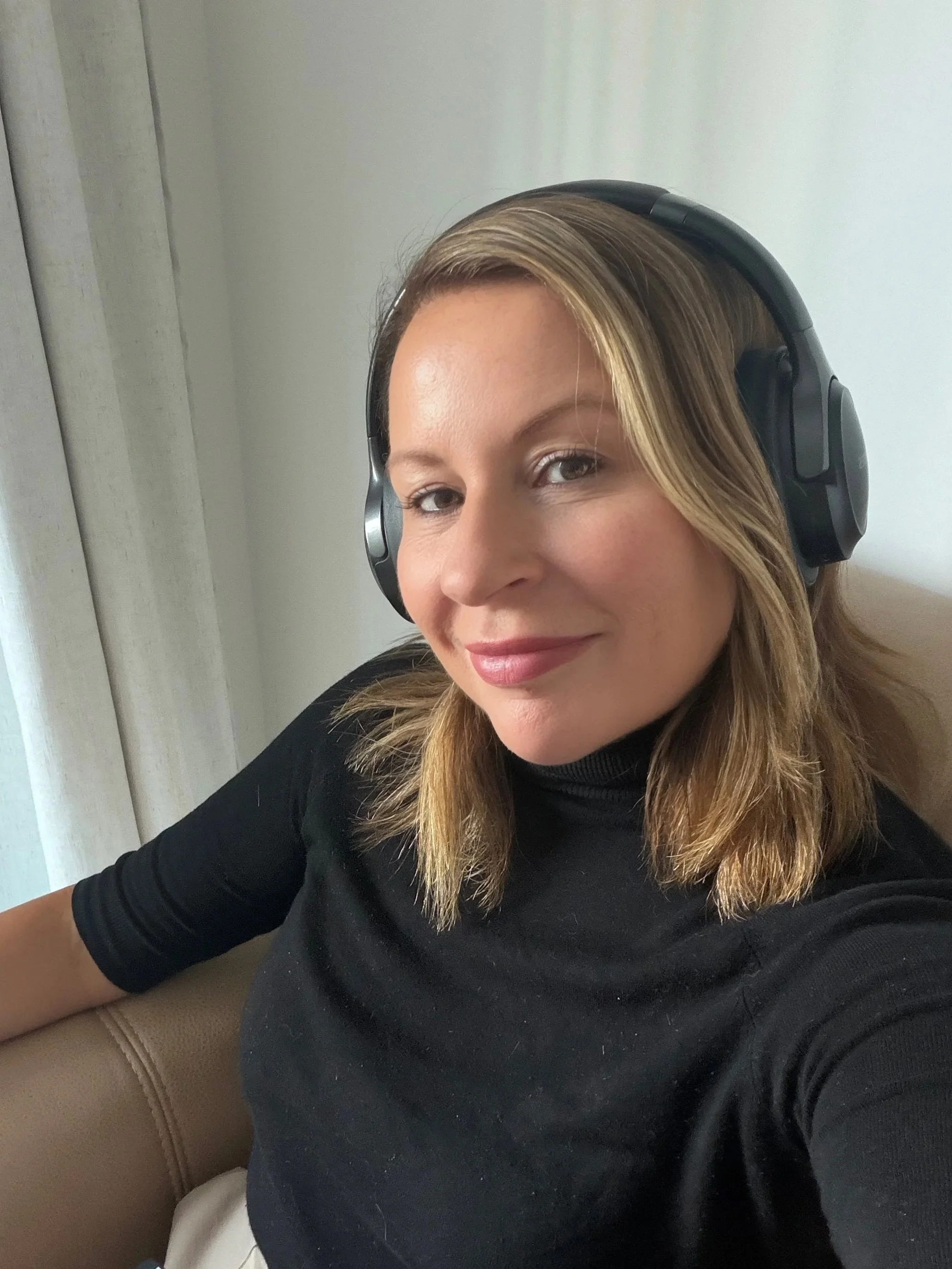A woman with blonde hair wearing a black long-sleeve shirt and black over-ear headphones, sitting on a tan-colored couch in a room with light-colored walls.