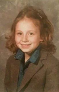 Young girl with curly red hair smiling, wearing a blazer and shirt.
