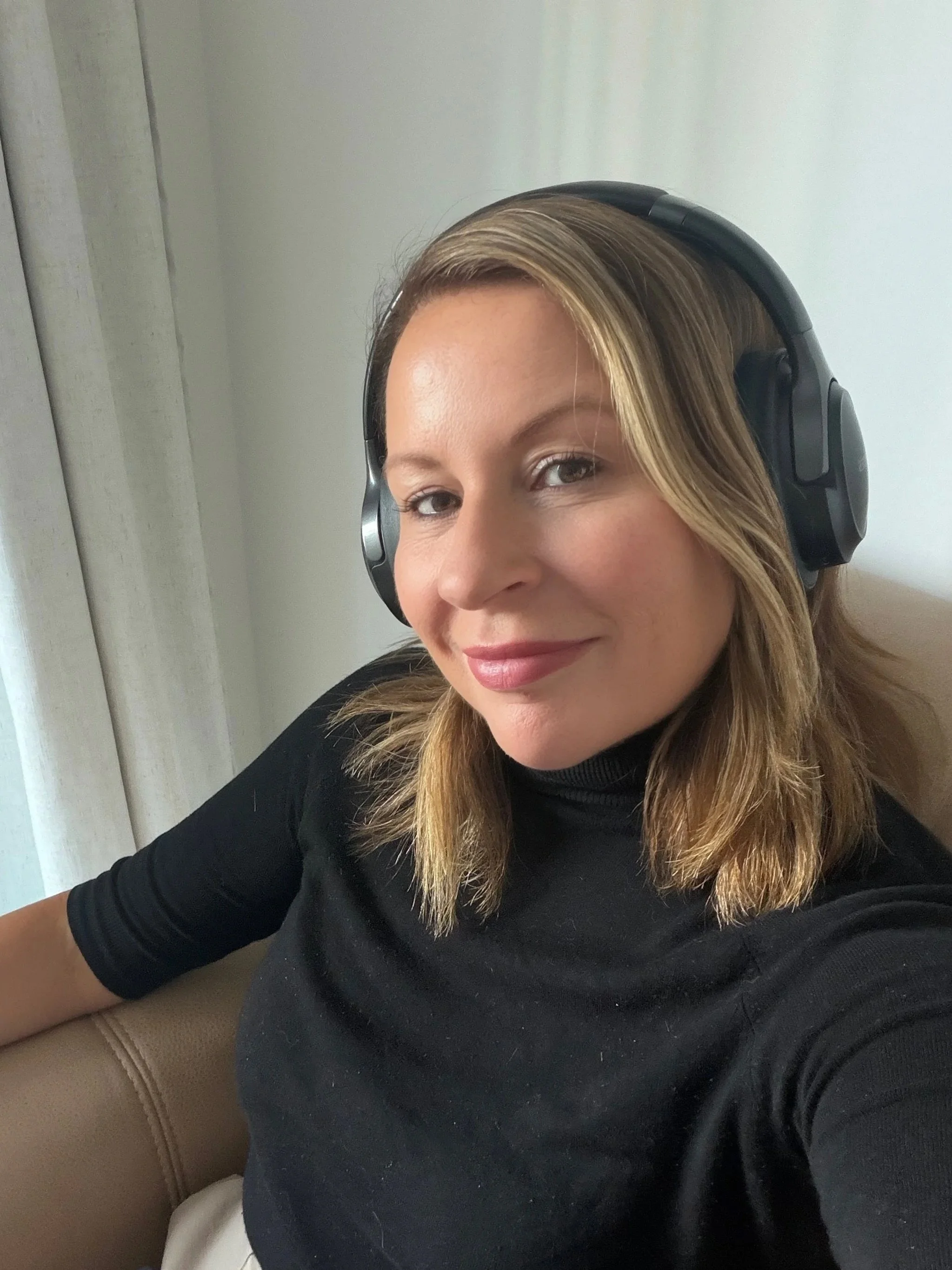 A woman with blonde, wavy hair wearing black headphones and a black shirt, sitting on a beige couch against a light-colored wall.