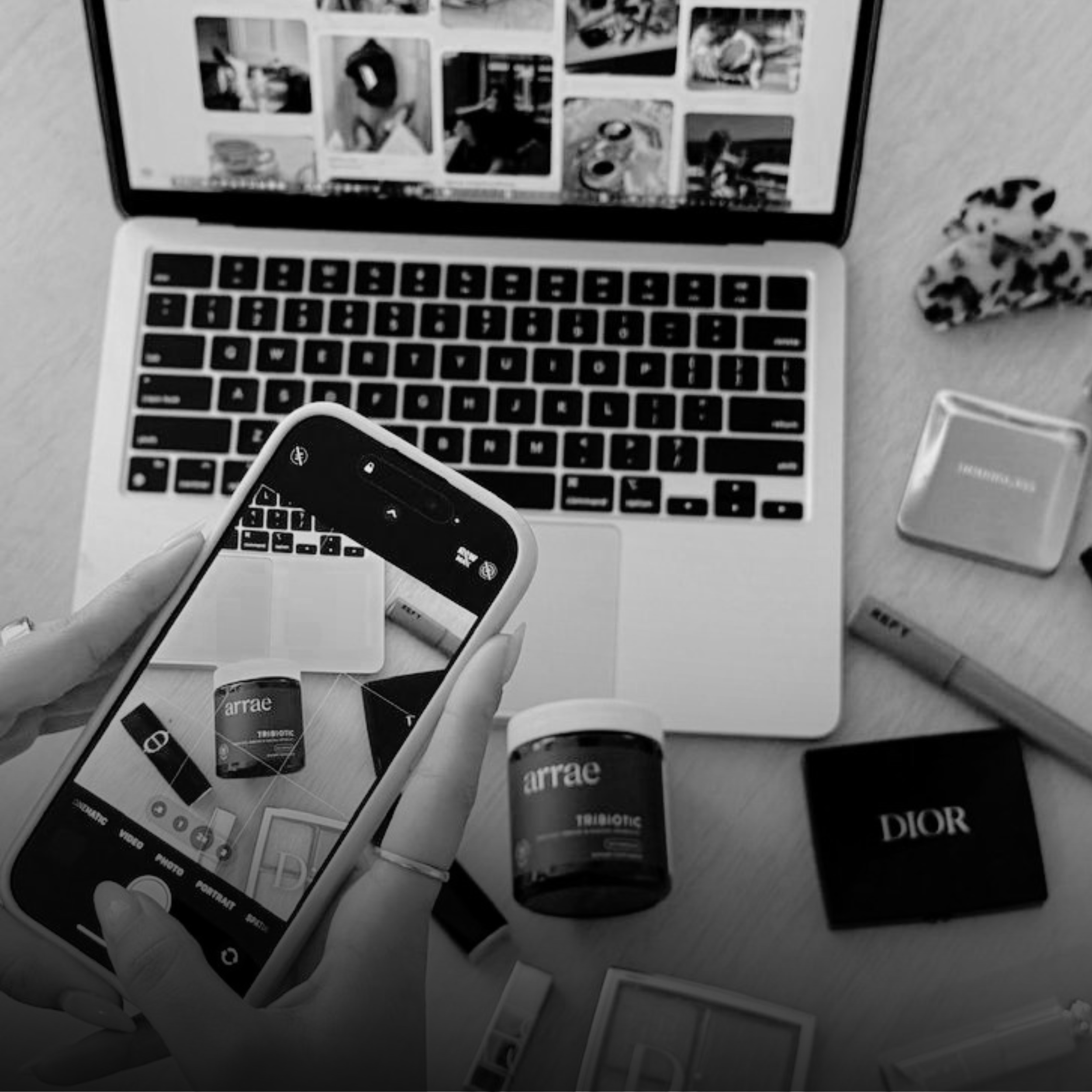 Black and white photo of a person holding a phone with the screen showing a camera app. The background includes a laptop with a collage of photos on the screen, a leopard print item, and various bottles and objects on the table.