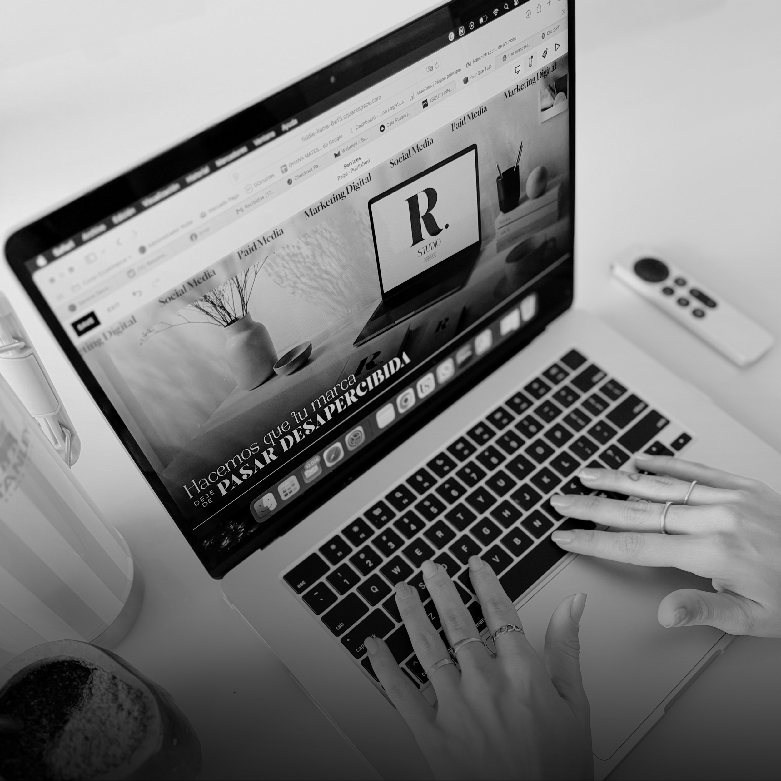 Black and white photograph of a person using a laptop with hands wearing rings, near a white mouse and a coffee mug, on a white surface.