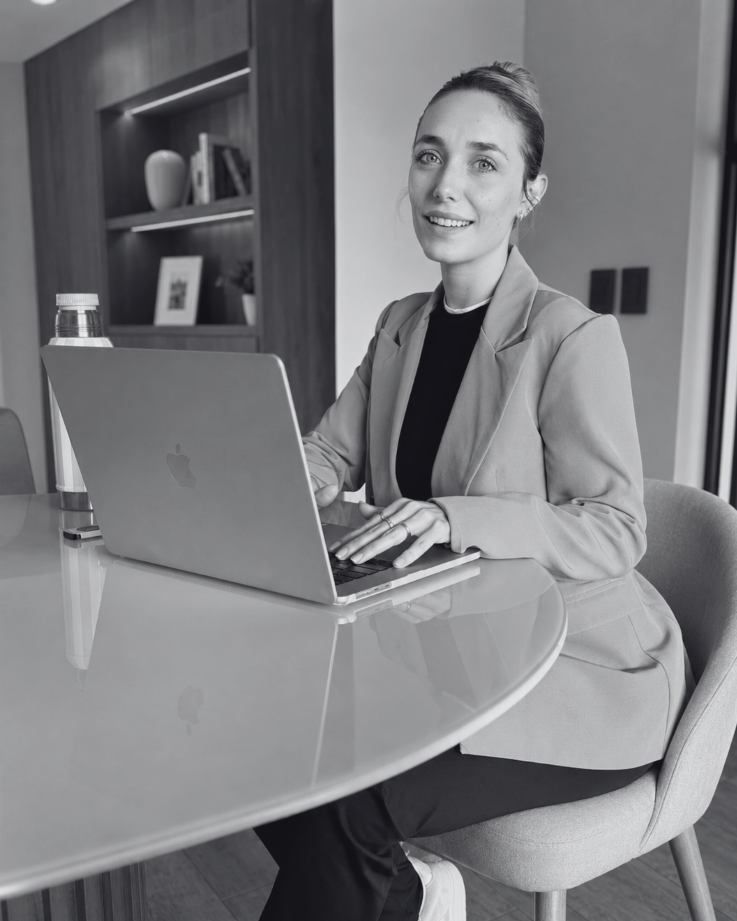 A woman with light skin and blonde hair sitting at a desk using a MacBook laptop, smiling at the camera, in a modern office setting.