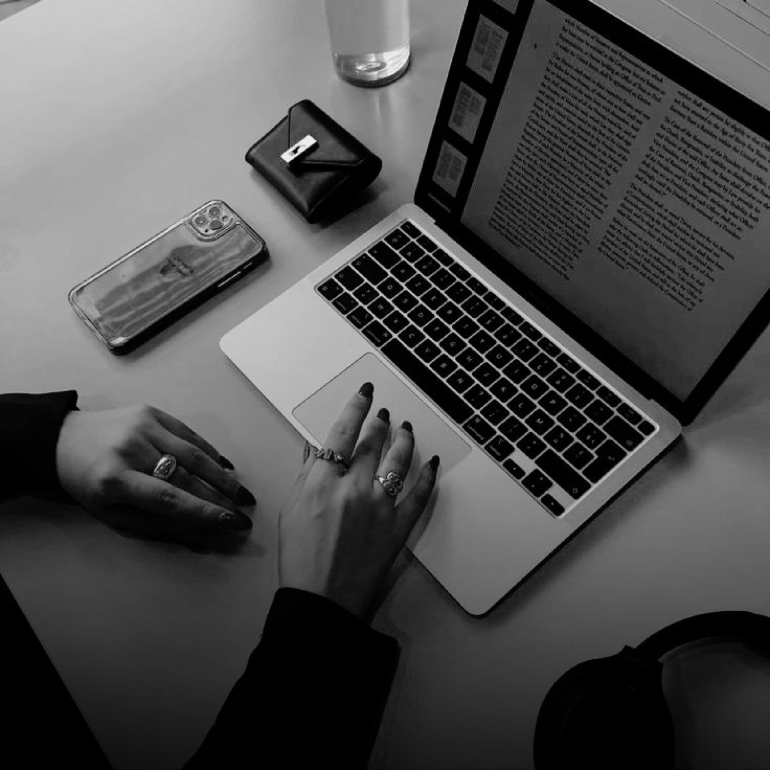 Black and white photo of a desk with a laptop showing text, a smartphone, a notebook with a pen, a bottle of water, and a hand with painted nails and rings.