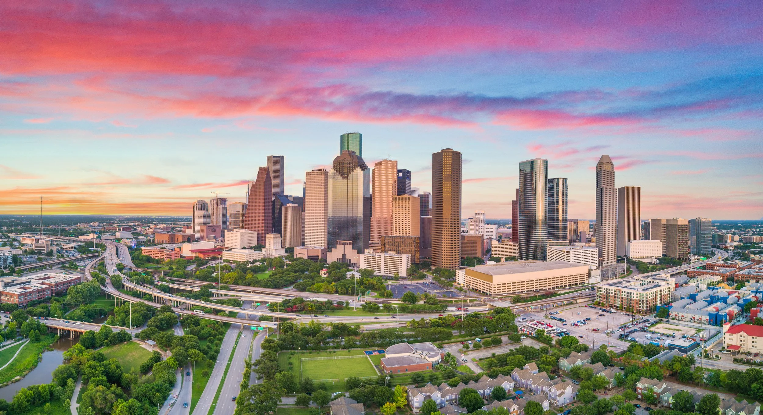 Aerial view of downtown Los Angeles at sunset with skyscrapers, highways, green parks, and a colorful sky.