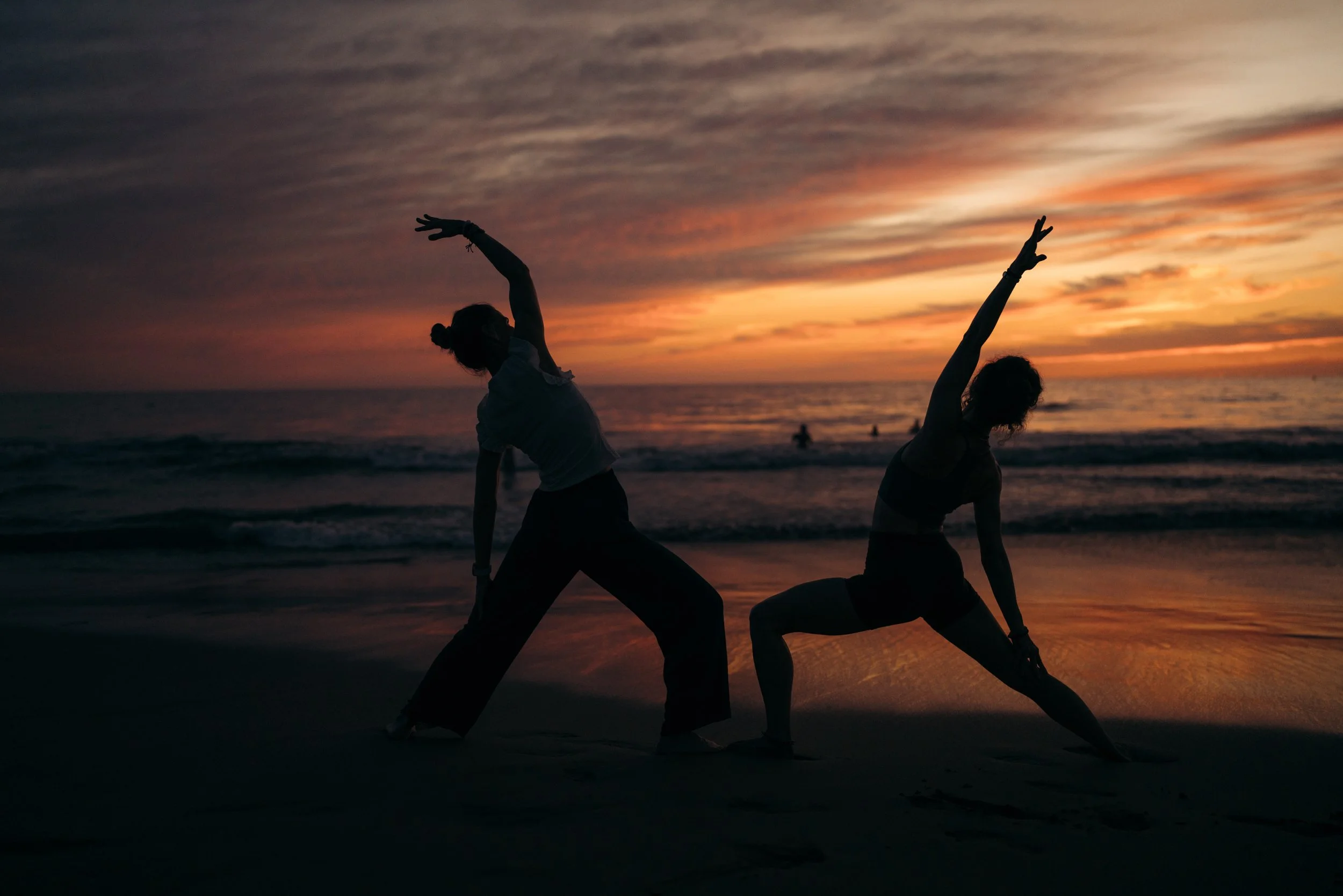 Two people practicing yoga on the beach during sunset, silhouetted against colorful sky.
