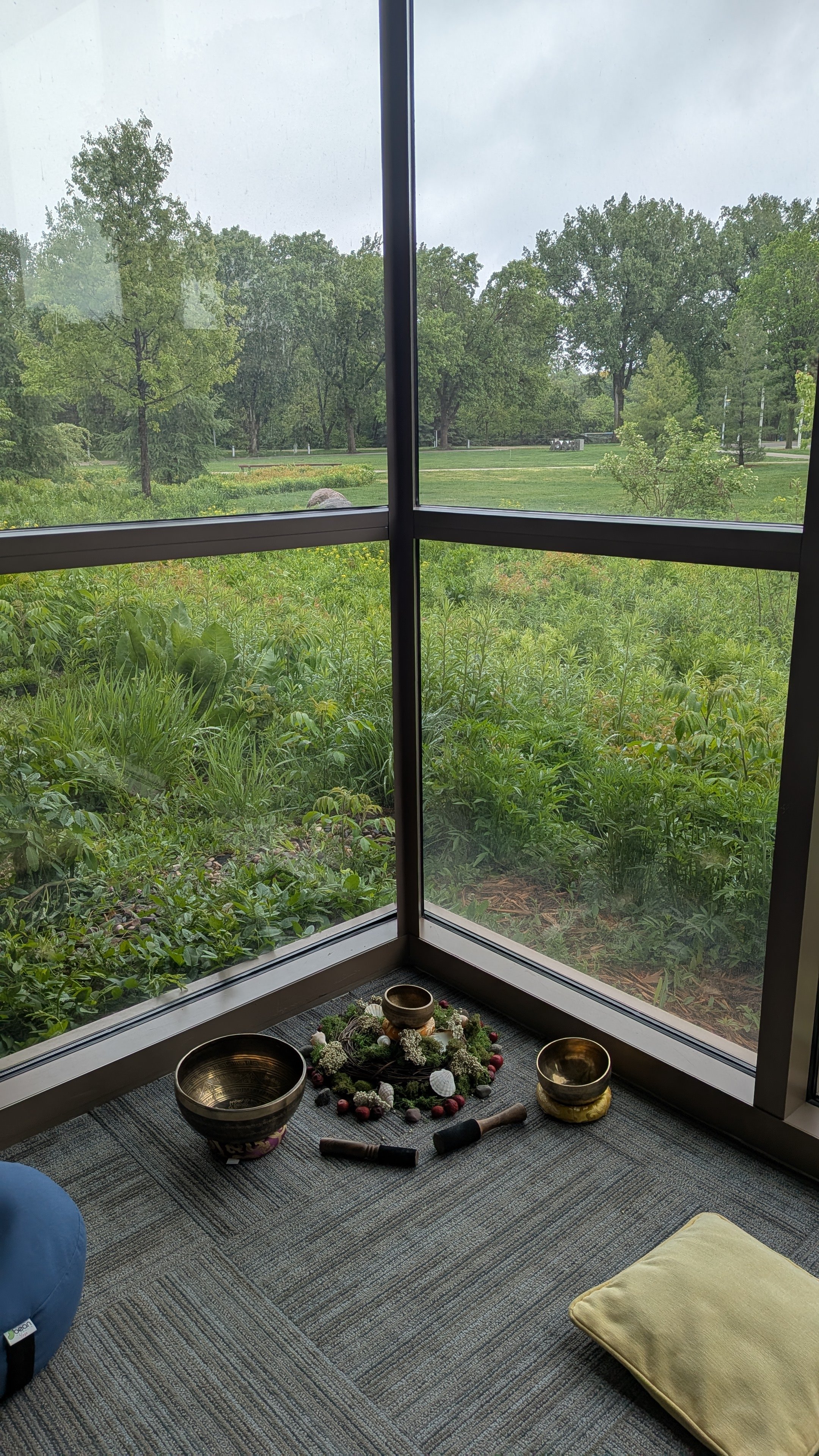 A corner room with a large window showing a green outdoor landscape with trees and grass. On the floor, there are singing bowls, a small mallet, a cushion, and a decorative arrangement of flowers and stones, suggesting a meditation or relaxation space.