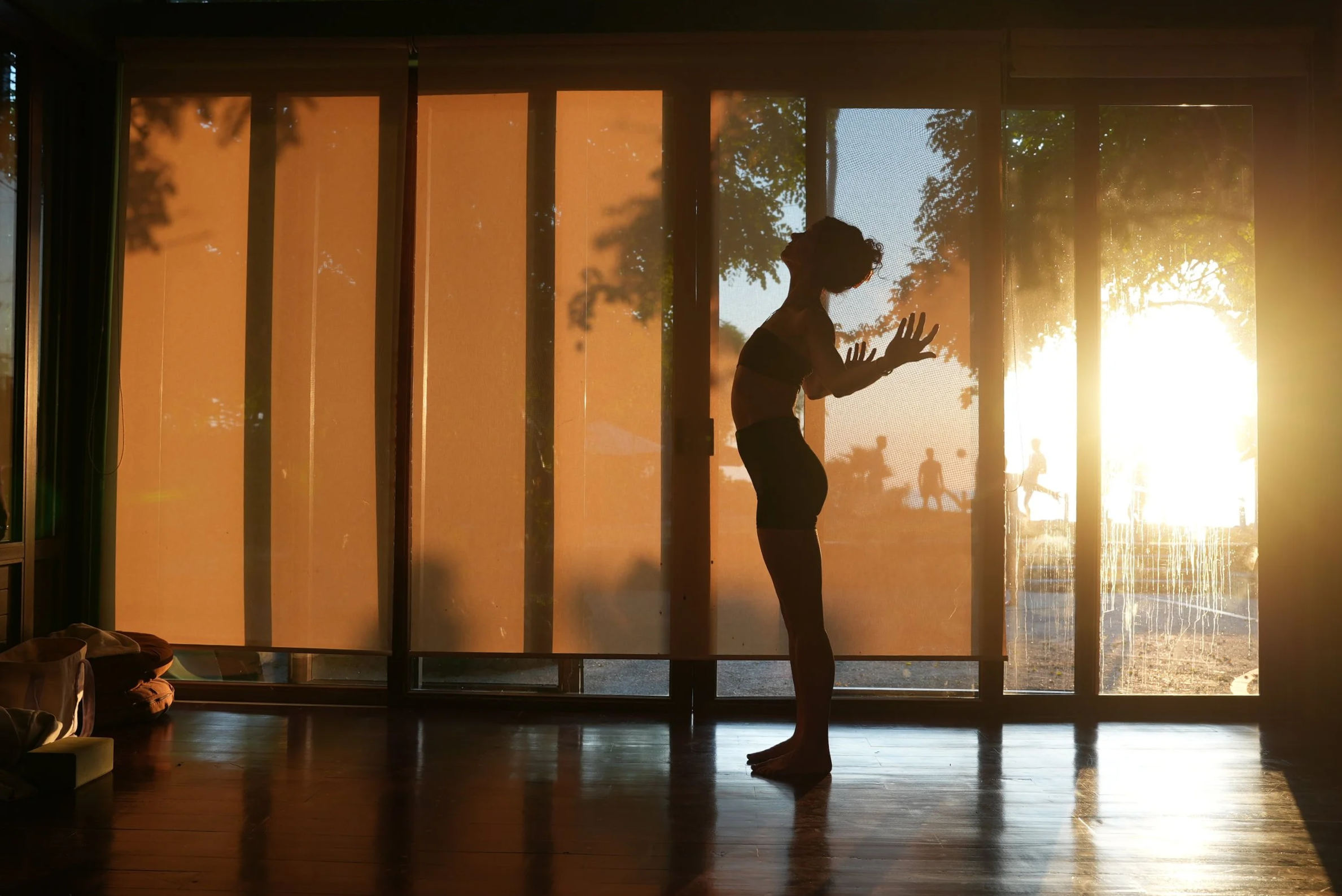 Silhouette of a person practicing yoga or stretching indoors near large windows at sunset, with trees and people outside.