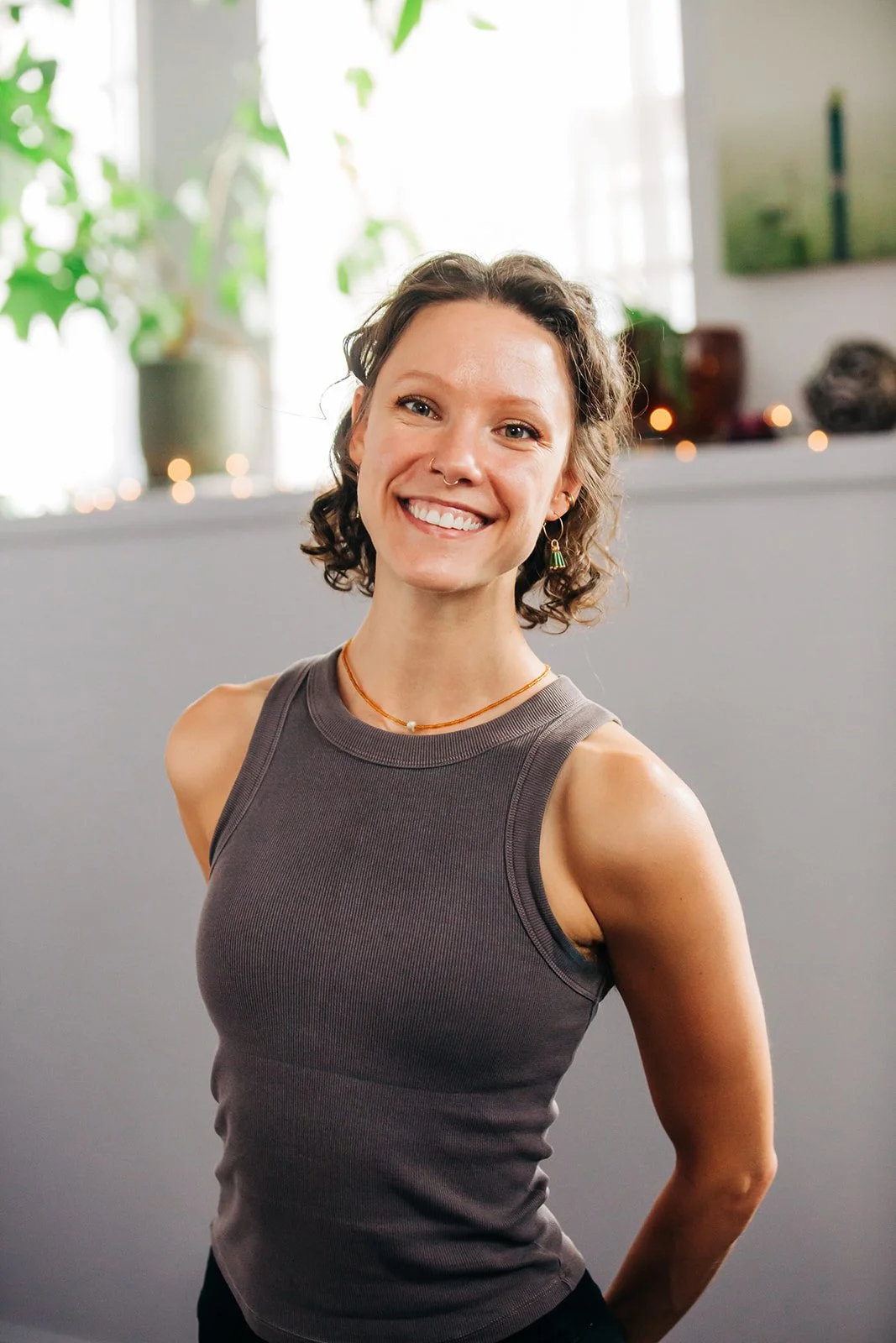 A smiling woman with curly brown hair wearing a sleeveless gray top standing indoors with a gray wall, decorative candles, and plants in the background.