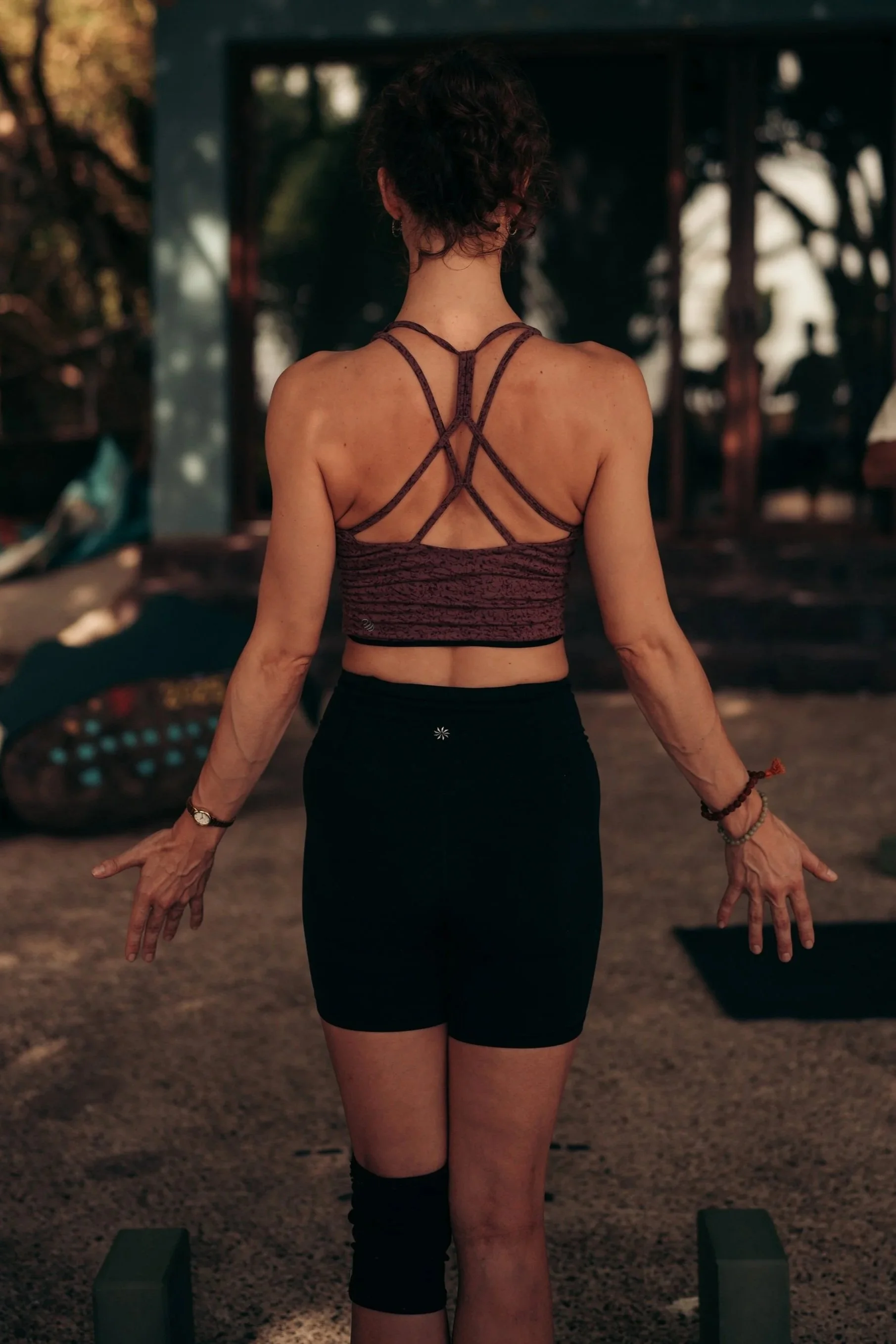A woman with short curly hair standing outdoors in a yoga pose, facing away from the camera, wearing a strappy sports bra and black shorts, and a knee brace, with her arms extended downward and hands relaxed.