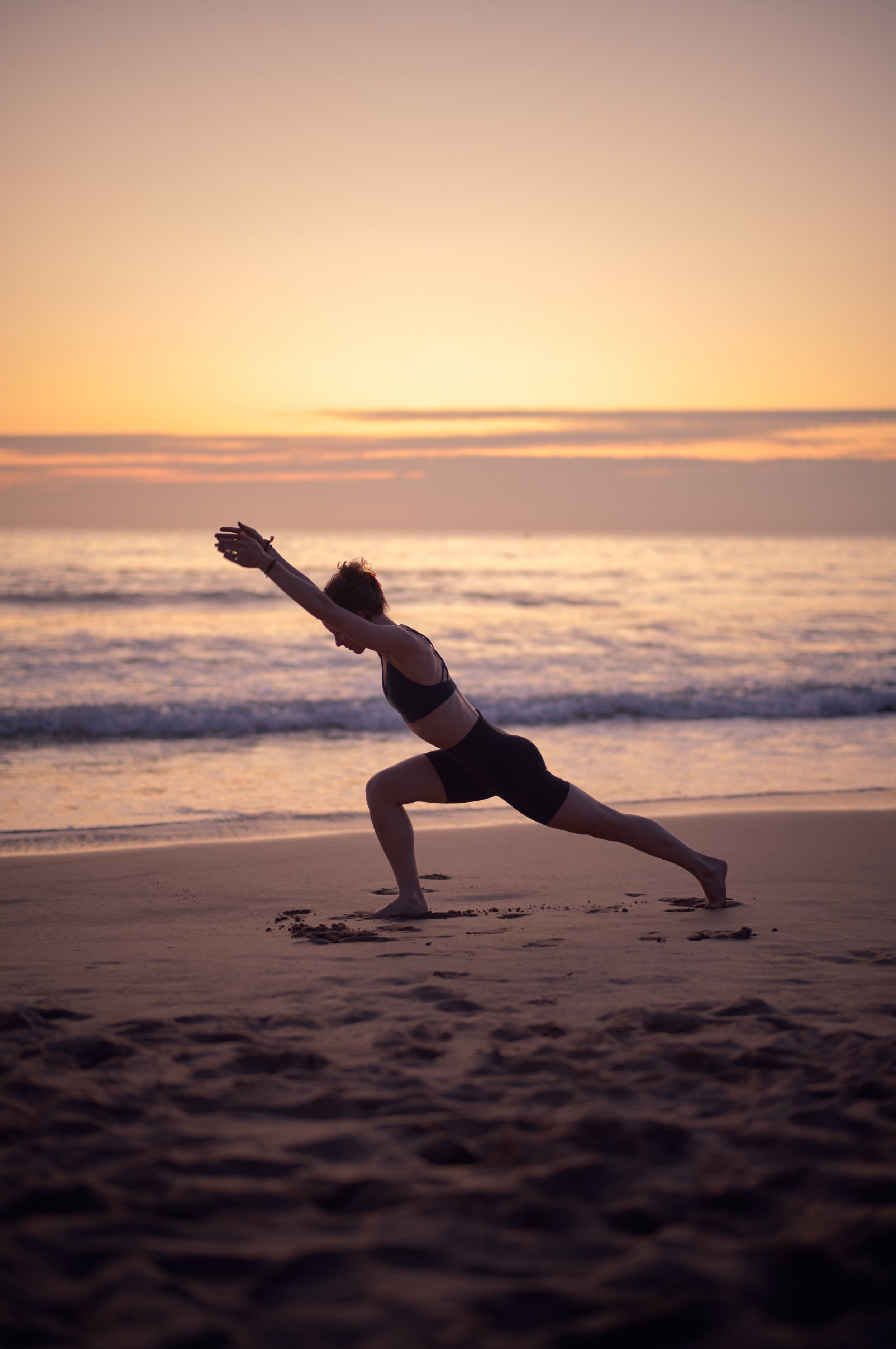 A woman practicing yoga on the beach at sunset, in a lunge pose with arms extended forward.