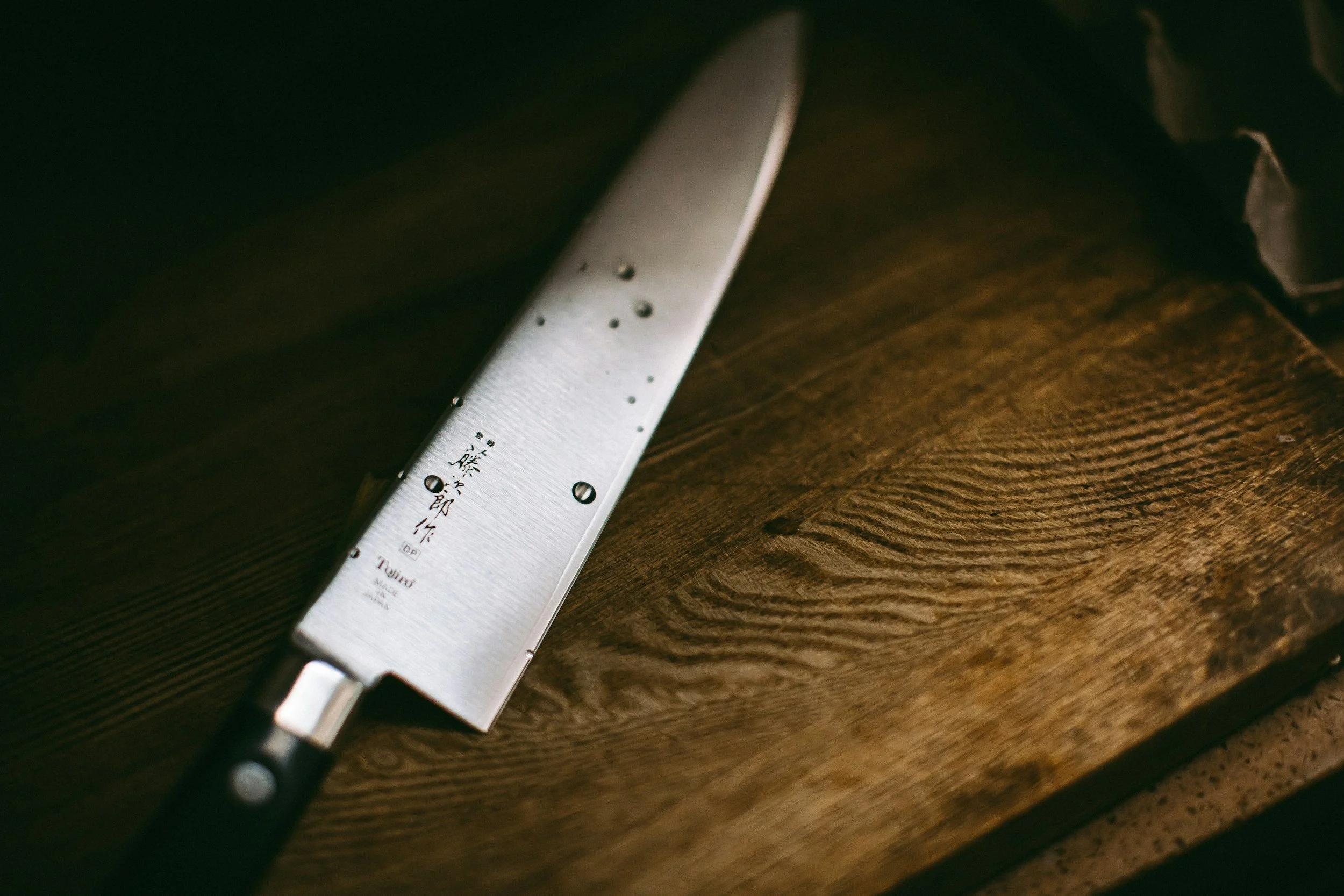 A close-up view of a metal knife with Japanese characters engraved on it, resting on a wooden surface.