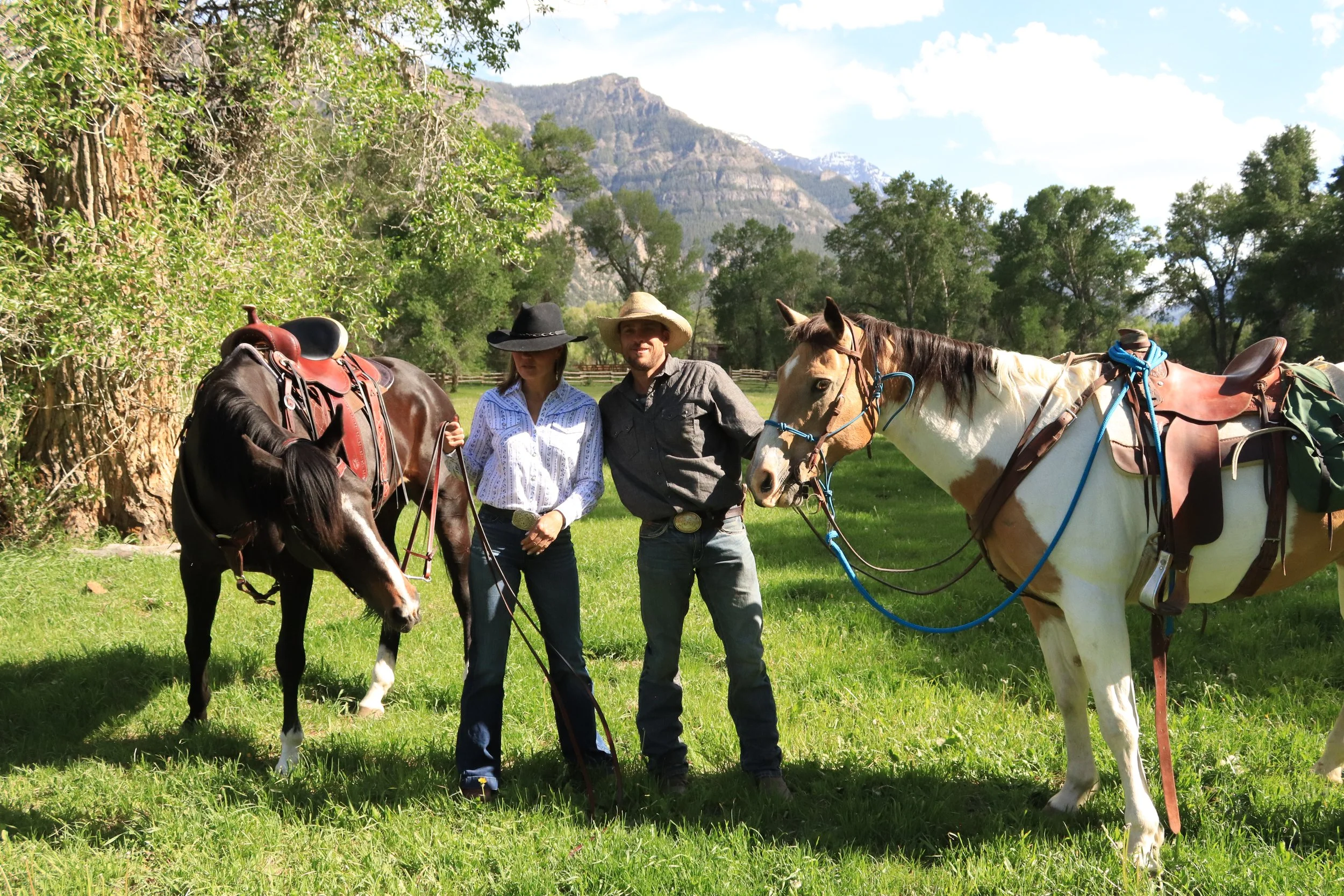 Two people standing outside with horses and mountain scenery in the background. The man and woman are smiling, and the horses have saddles on them.