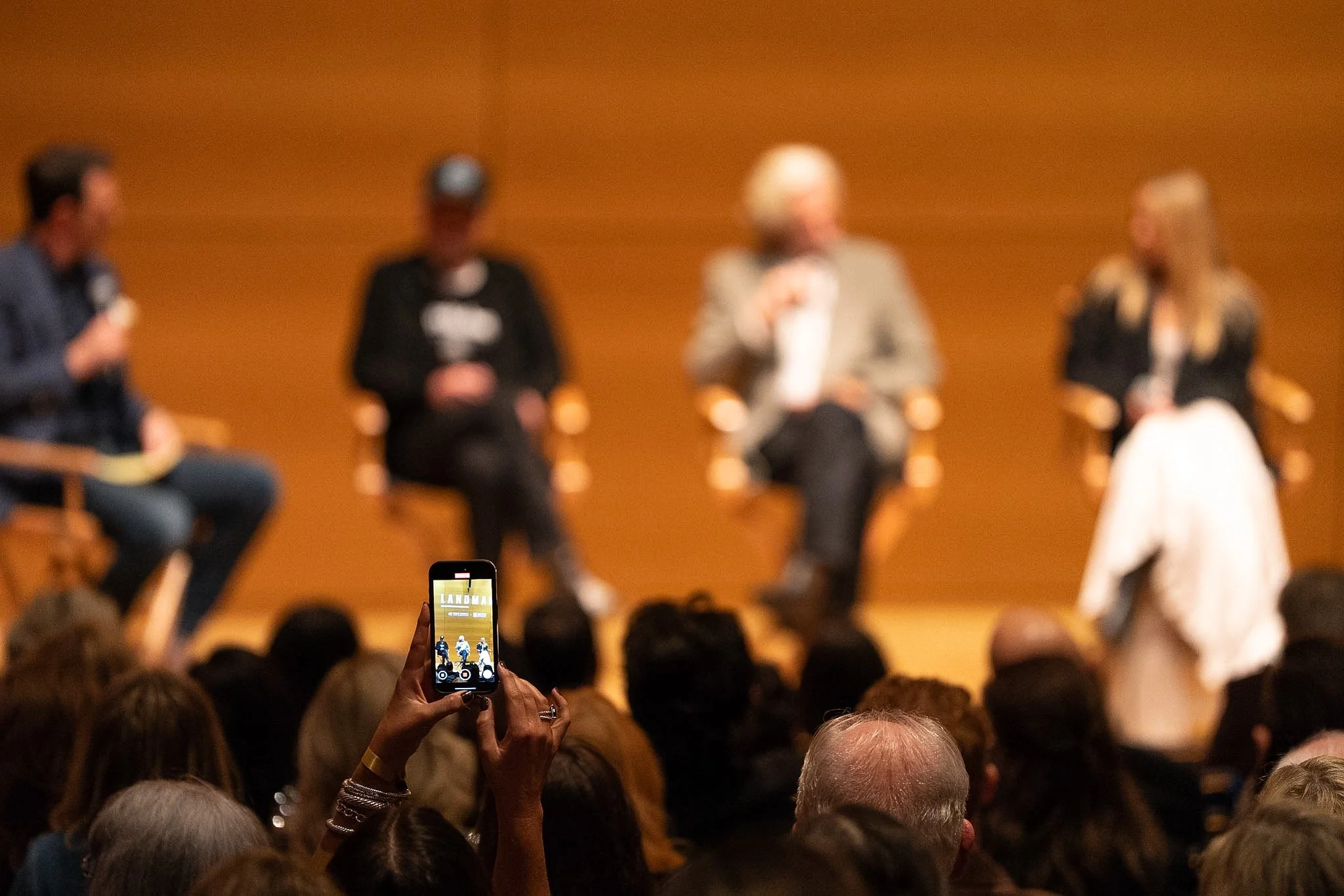 A panel discussion with four people seated in front of an audience, one person in the audience is taking a photo with their phone.