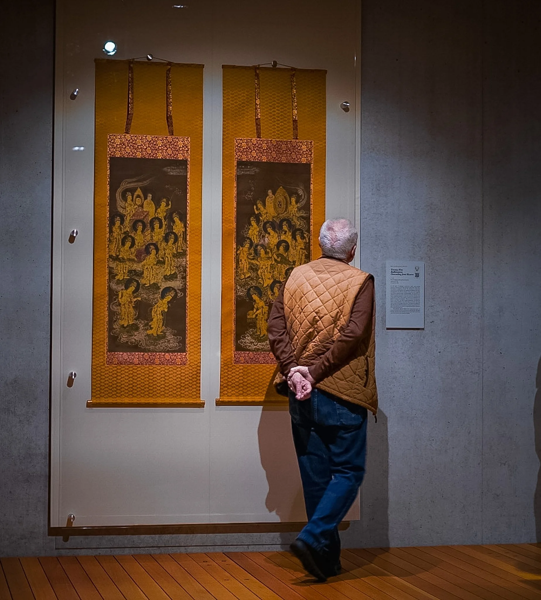An elderly man with gray hair, wearing a tan quilted vest over a shirt, blue jeans, and black shoes, stands in front of two hanging ornate tapestries with gold and black designs inside an art gallery. The man is looking up at the tapestries, which ar