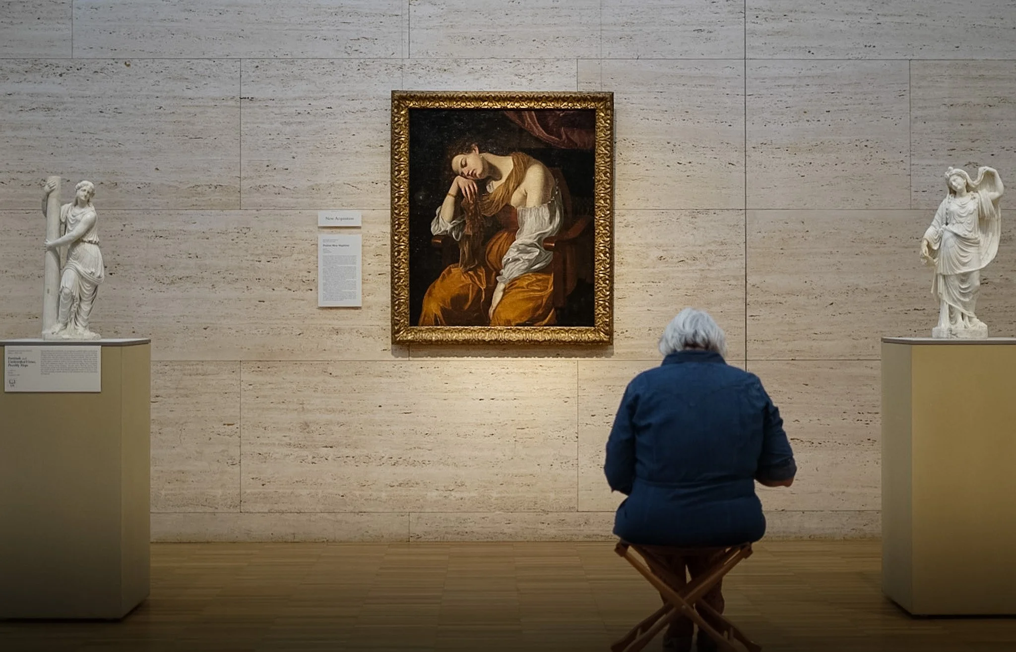 A person with gray hair sitting on a foldable chair in front of a painting of a woman resting her head on her hand in an art gallery, with two classical statues on pedestals on either side.