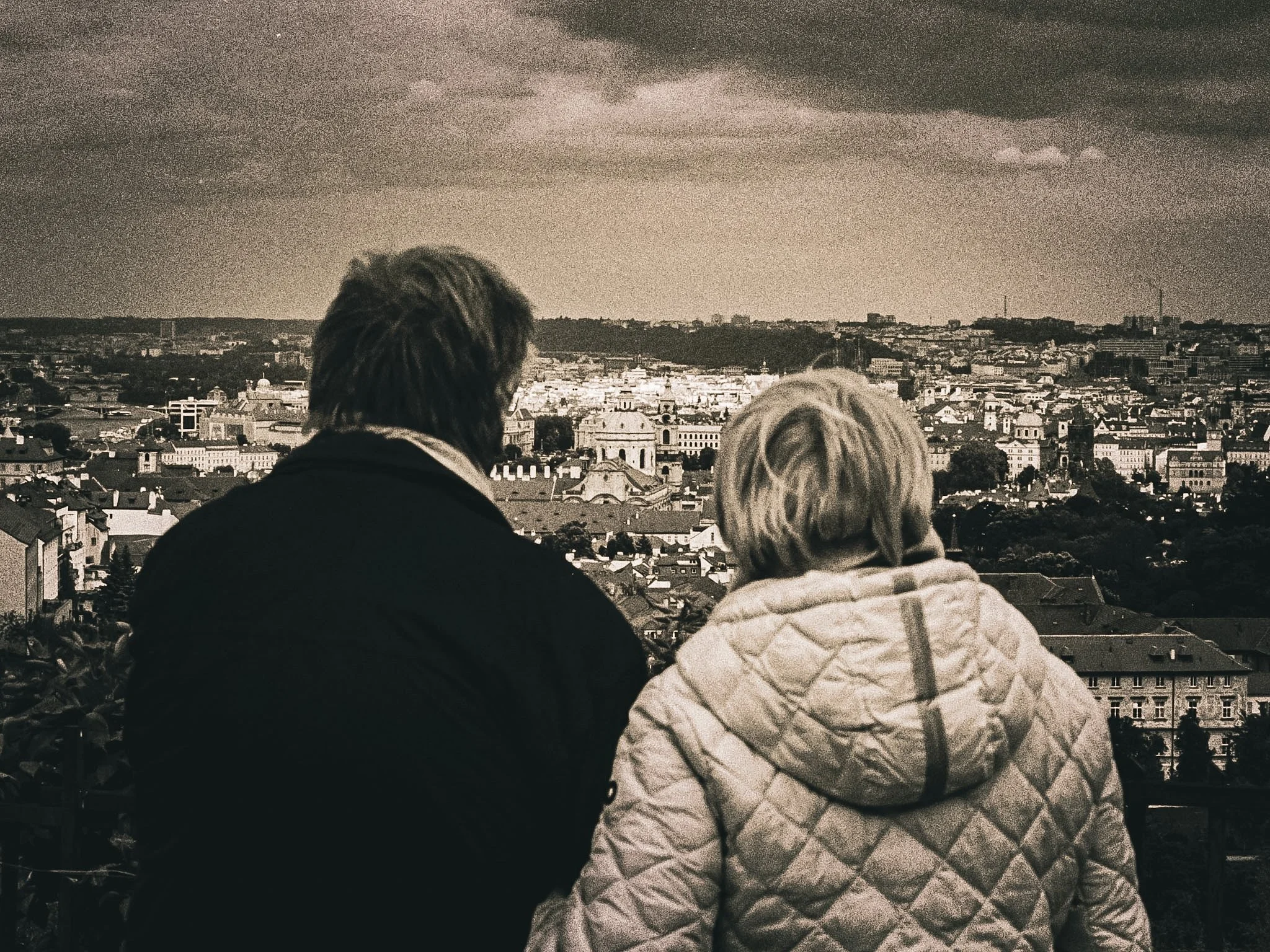 Two people sitting and overlooking a cityscape with historic buildings, under a cloudy sky.