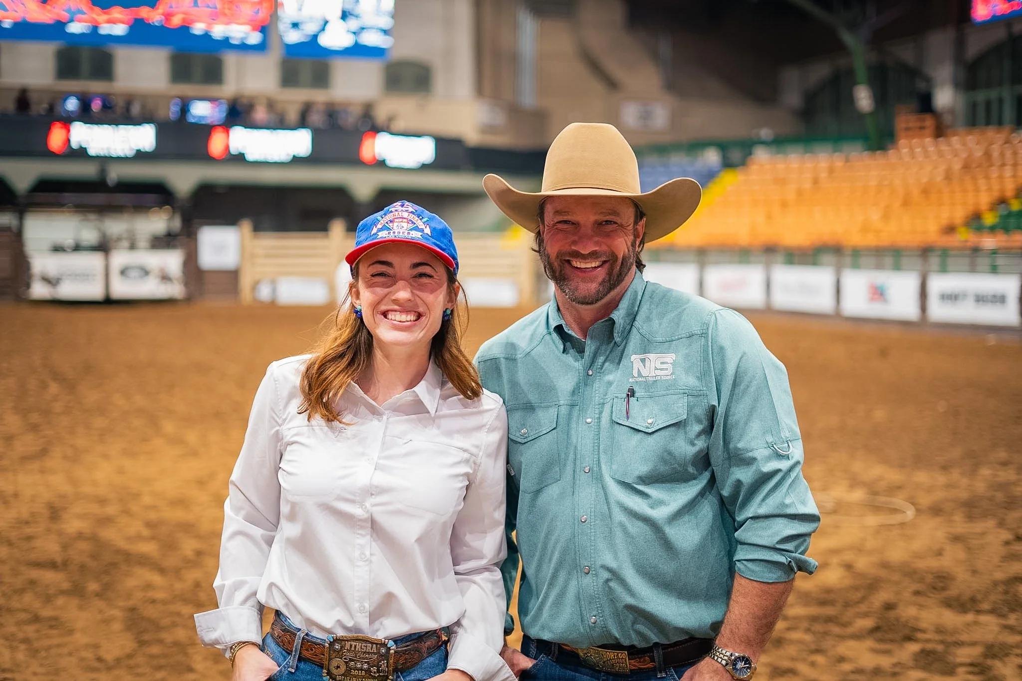 A woman and a man standing together at an indoor arena (Cowtown Coliseum), both smiling, with the woman wearing a blue cap and a white shirt, and the man wearing a cowboy hat and a light blue shirt.
