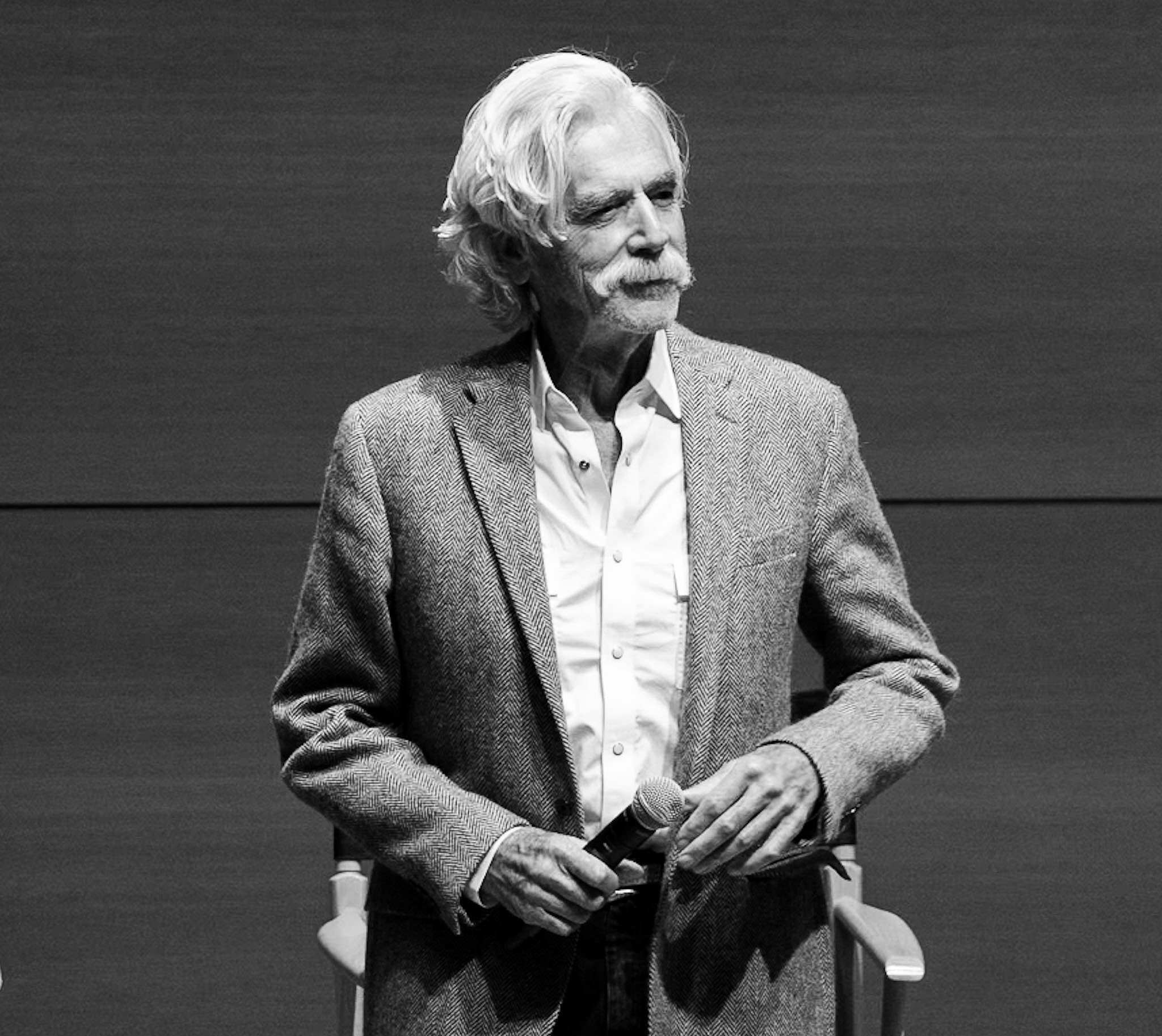 A black and white image of Sam Elliot with white hair and a mustache, dressed in a patterned blazer and white shirt, standing next to a microphone on a stage or panel at Landman's Premier in Fort Worth.