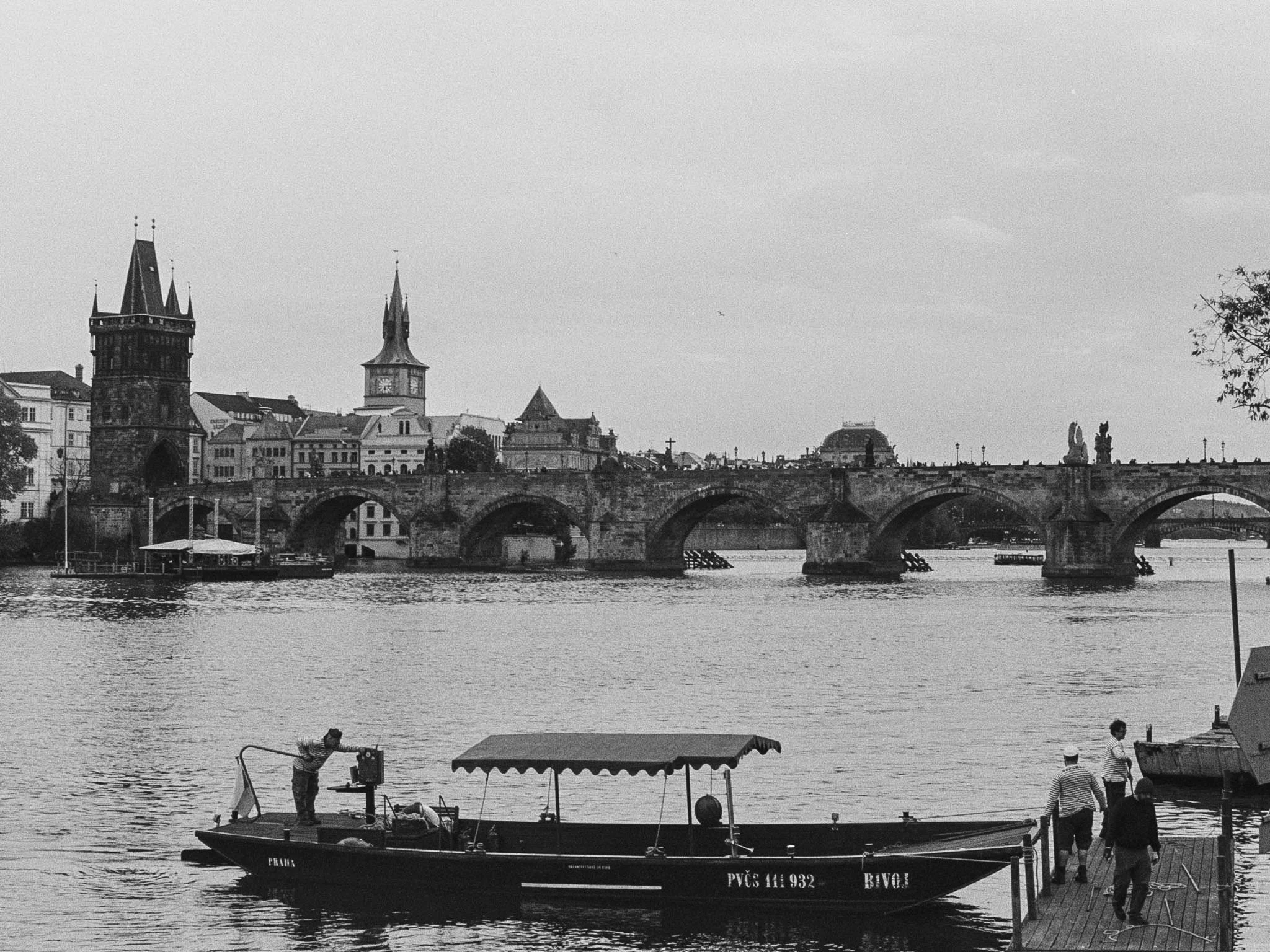 A black and white photo of a city skyline with historic buildings, a bridge crossing a river, and boats docked at the waterfront.
